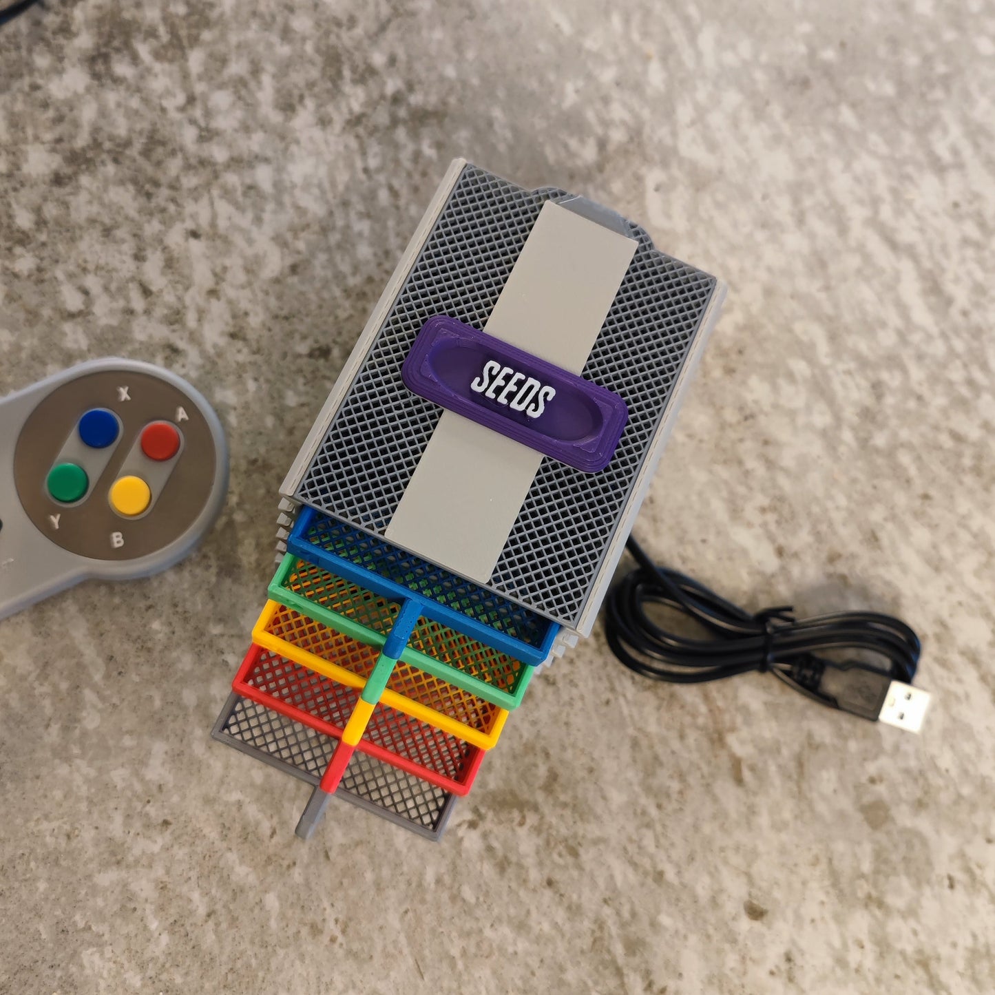 The Seed Dryer - Super Classic Console by PepperMerchant.net includes a retro game console-style "SEEDS" container with colorful trays, a USB-powered seed dryer, and a controller with red, yellow, green, and blue buttons on a gray surface.