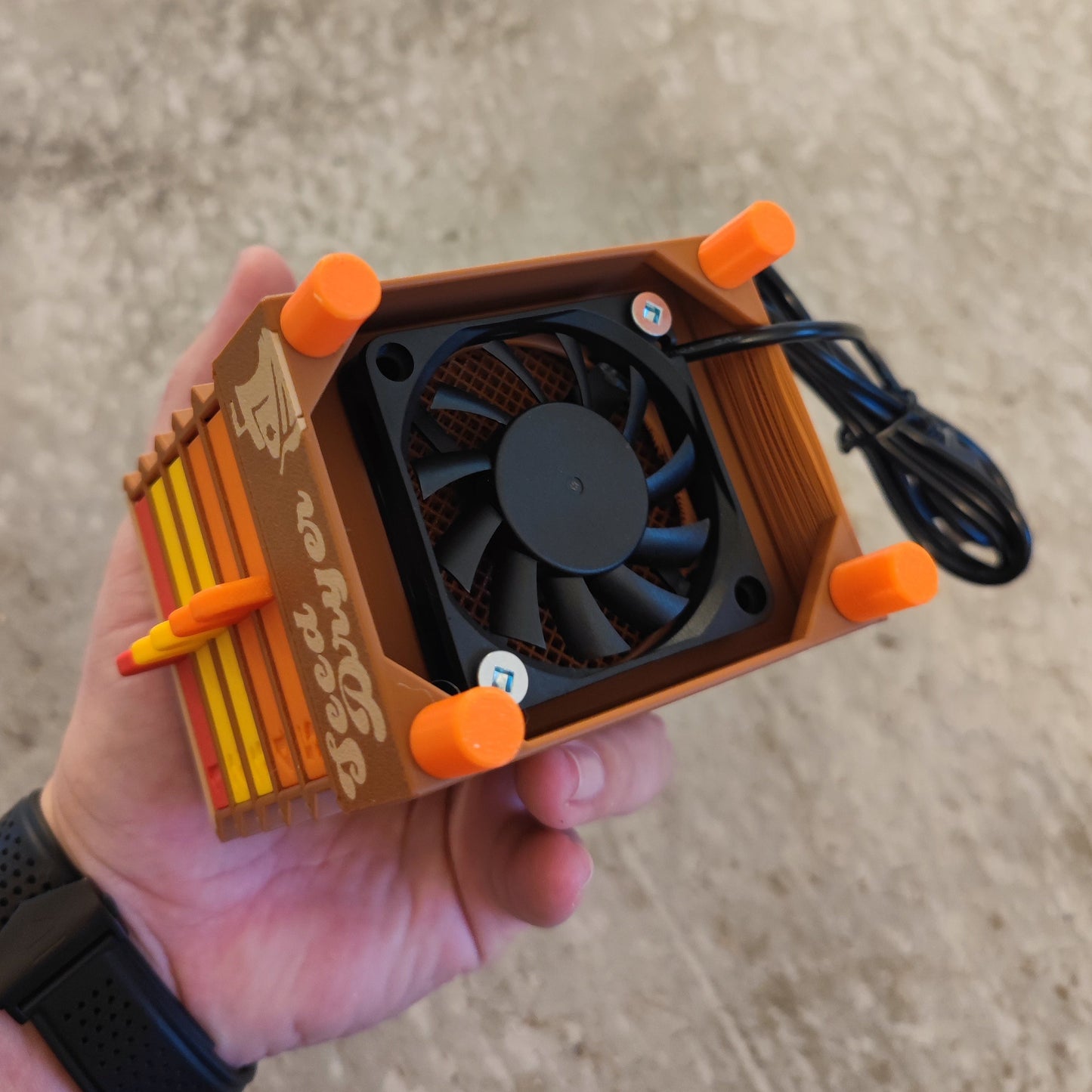 A hand holds a small, orange and brown Seed Dryer - Harvest Time by PepperMerchant.net. It features a black cooling fan, orange corner supports, a black wire, and a retro case design with branding on the side.