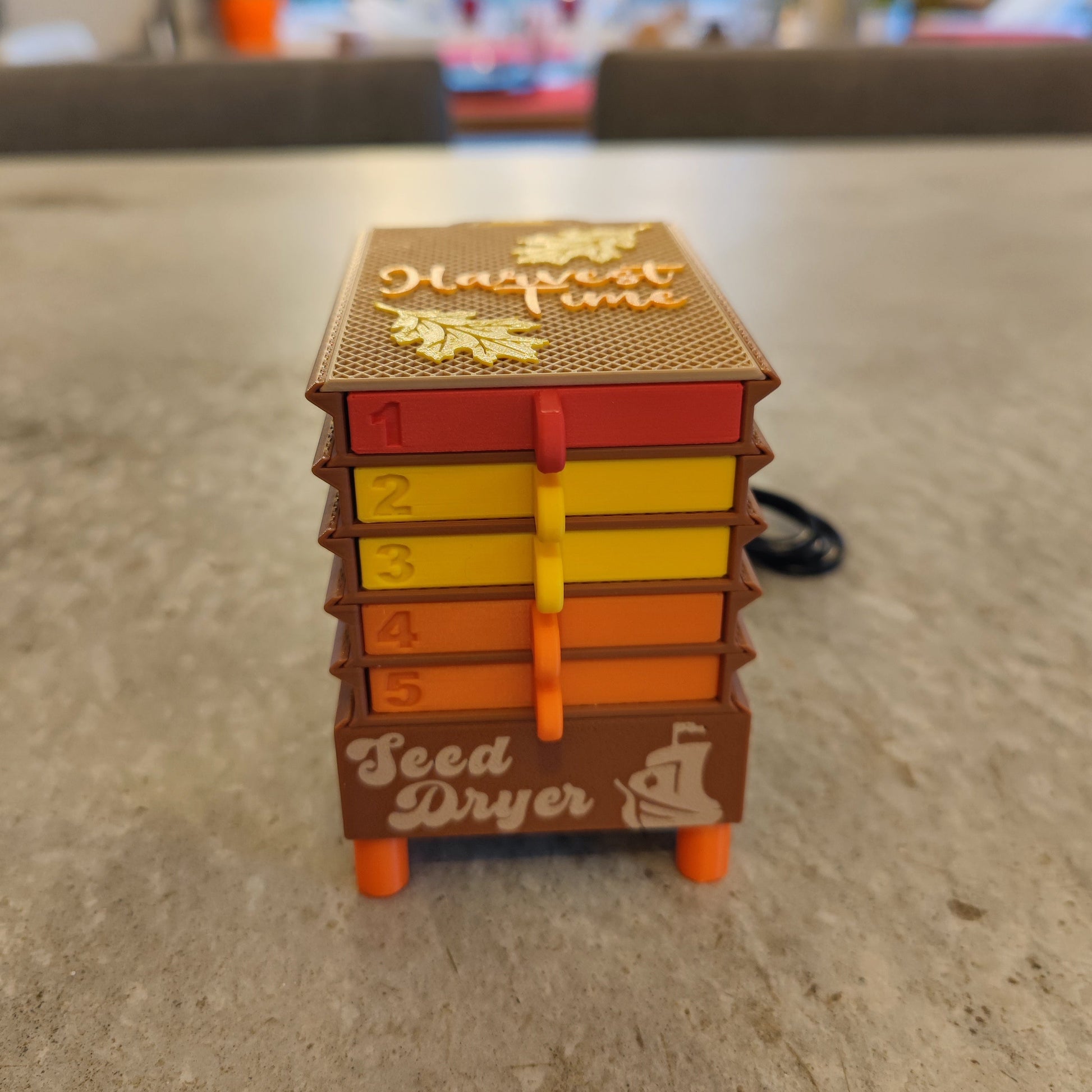 The Seed Dryer - Harvest Time by PepperMerchant.net is a miniature model with five colorful, numbered drawers and labeled front—ideal for demonstrating seed saving methods on any gray surface.