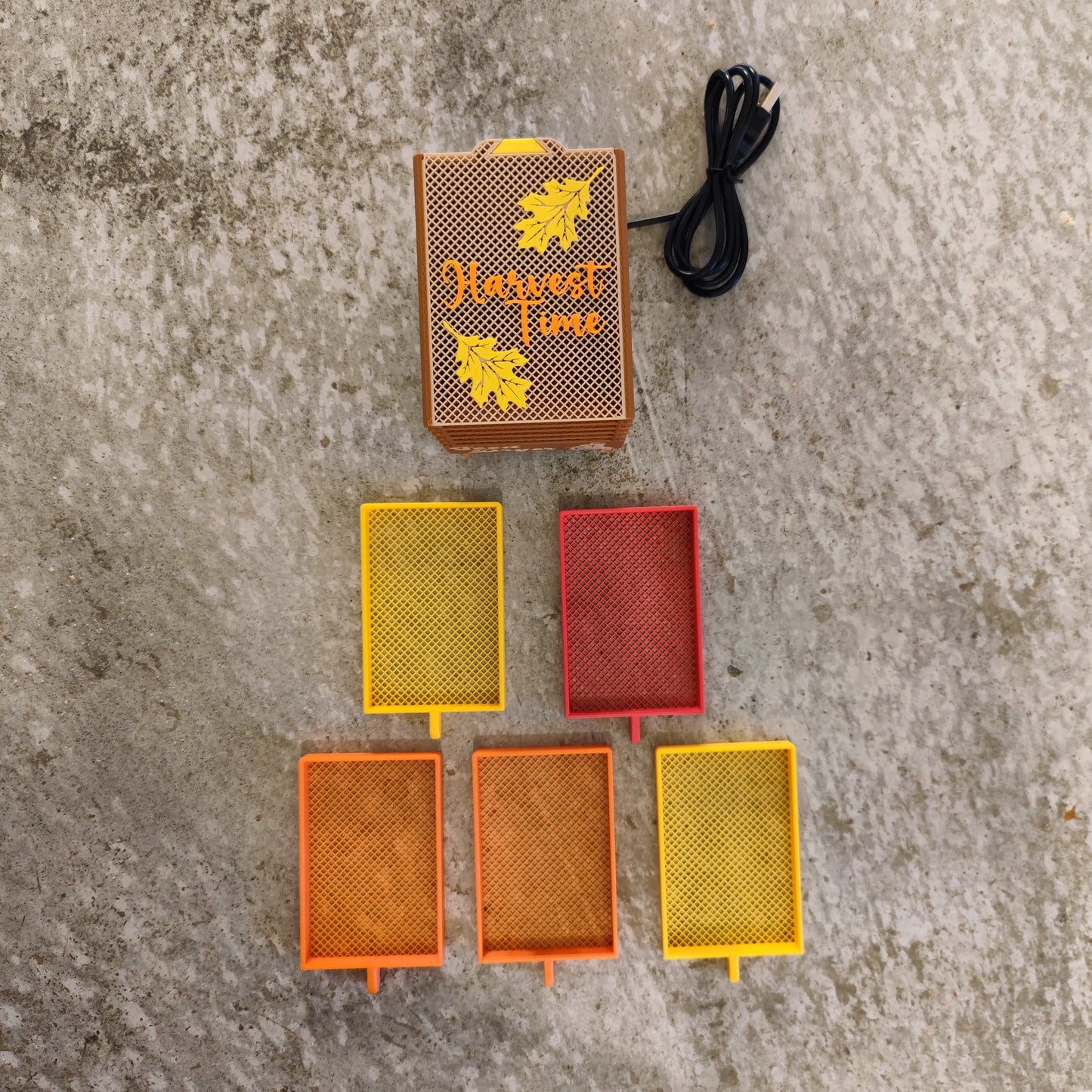 The Seed Dryer - Harvest Time by PepperMerchant.net features five colorful mesh trays, a black cord, and a brown box with leaf designs—offering a heatless way to save seeds—displayed on concrete.