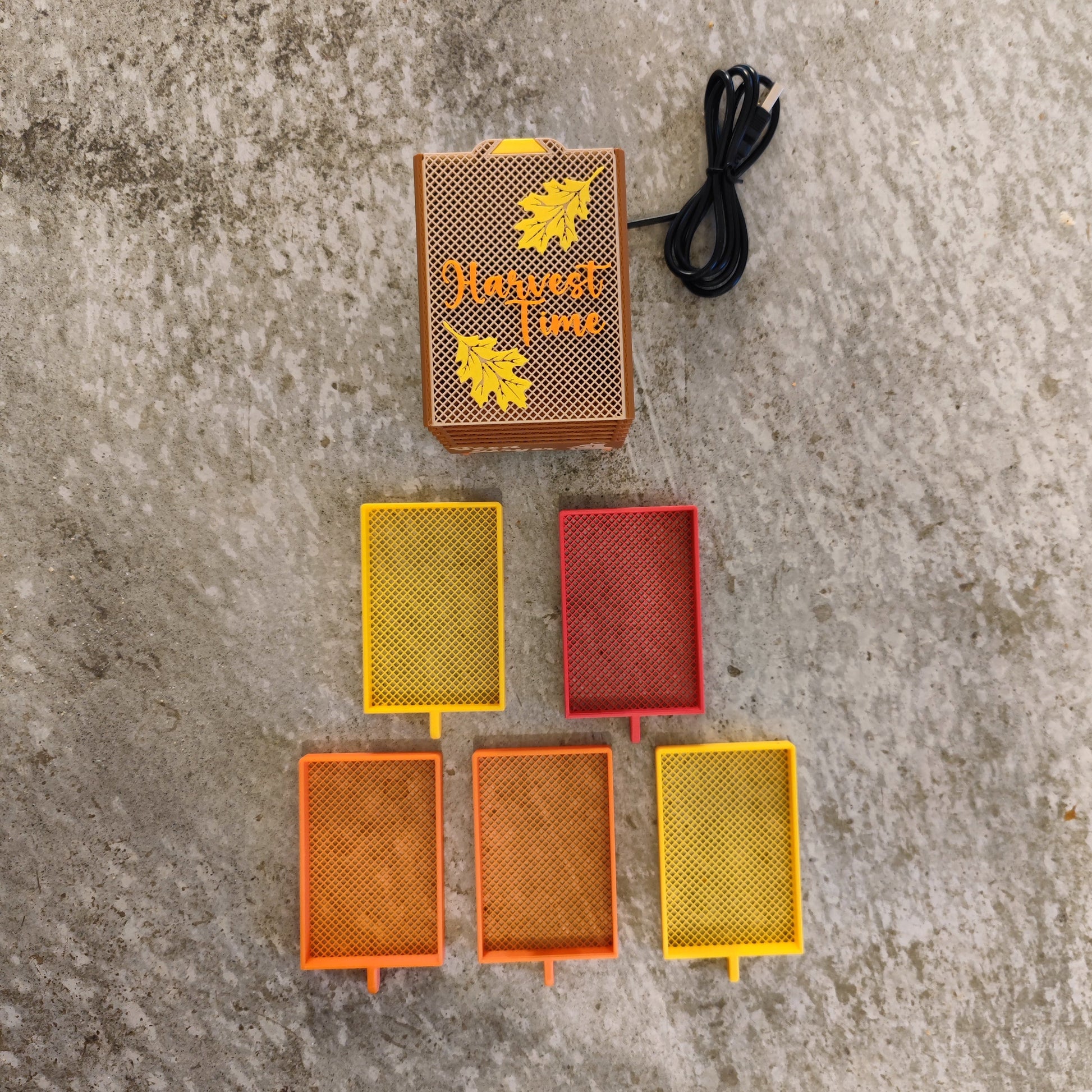 The Seed Dryer - Harvest Time by PepperMerchant.net features five colorful mesh trays, a black cord, and a brown box with leaf designs—offering a heatless way to save seeds—displayed on concrete.