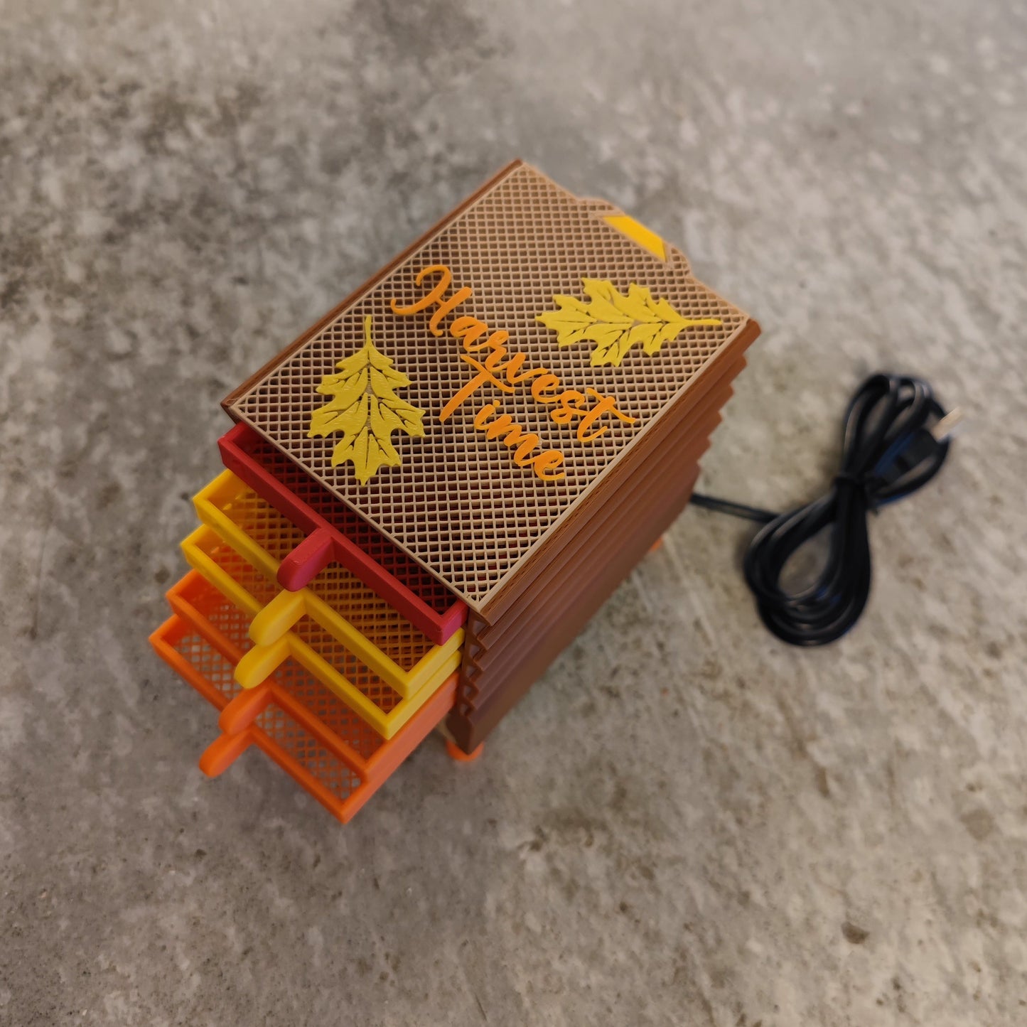 The Seed Dryer - Harvest Time by PepperMerchant.net, with autumn leaf lid and orange-yellow stacked trays, rests on a textured gray surface with its coiled black power cord—ideal for efficient seed saving.