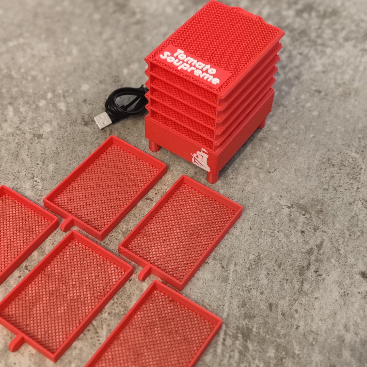 A stack of red Seed Dryer - Tomato Soupreme trays from PepperMerchant.net sits on a gray surface with six tray inserts and a USB-powered seed dryer, demonstrating modern seed-saving technology.