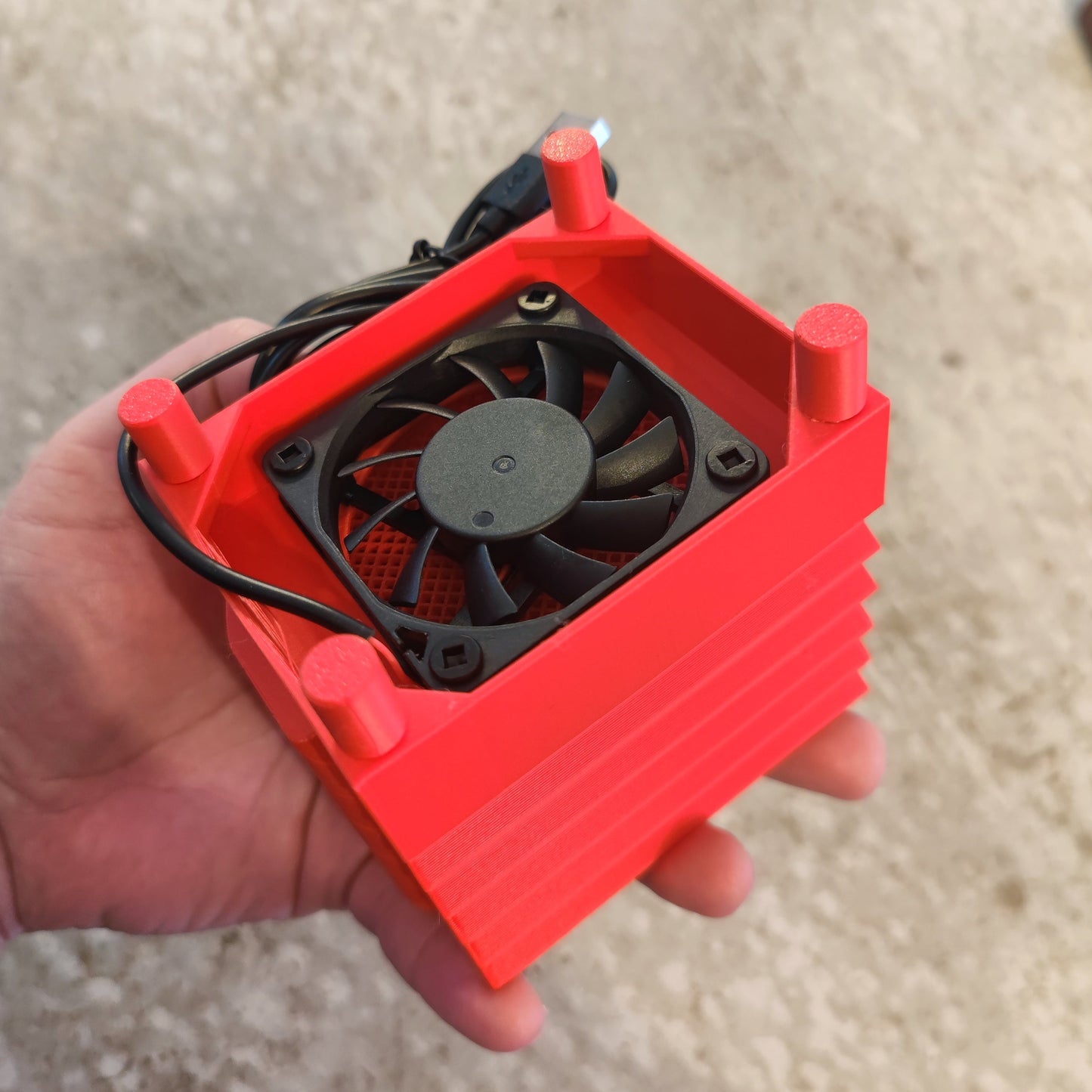 A hand holds the PepperMerchant.net Seed Dryer - Tomato Soupreme, a compact red USB-powered device with a black cooling fan and visible side fins, attached to a USB cable, set against a blurred light background.