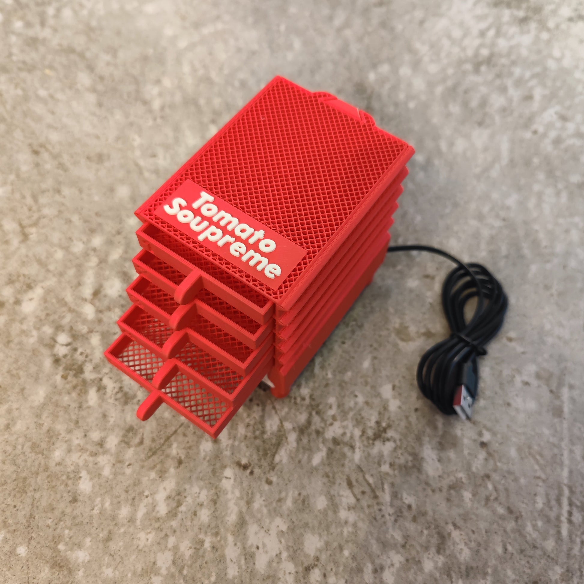 The PepperMerchant.net Seed Dryer - Tomato Soupreme is a compact, red, rectangular USB-powered seed dryer with a mesh texture and vented layers, shown on a speckled gray surface with its USB cable attached.
