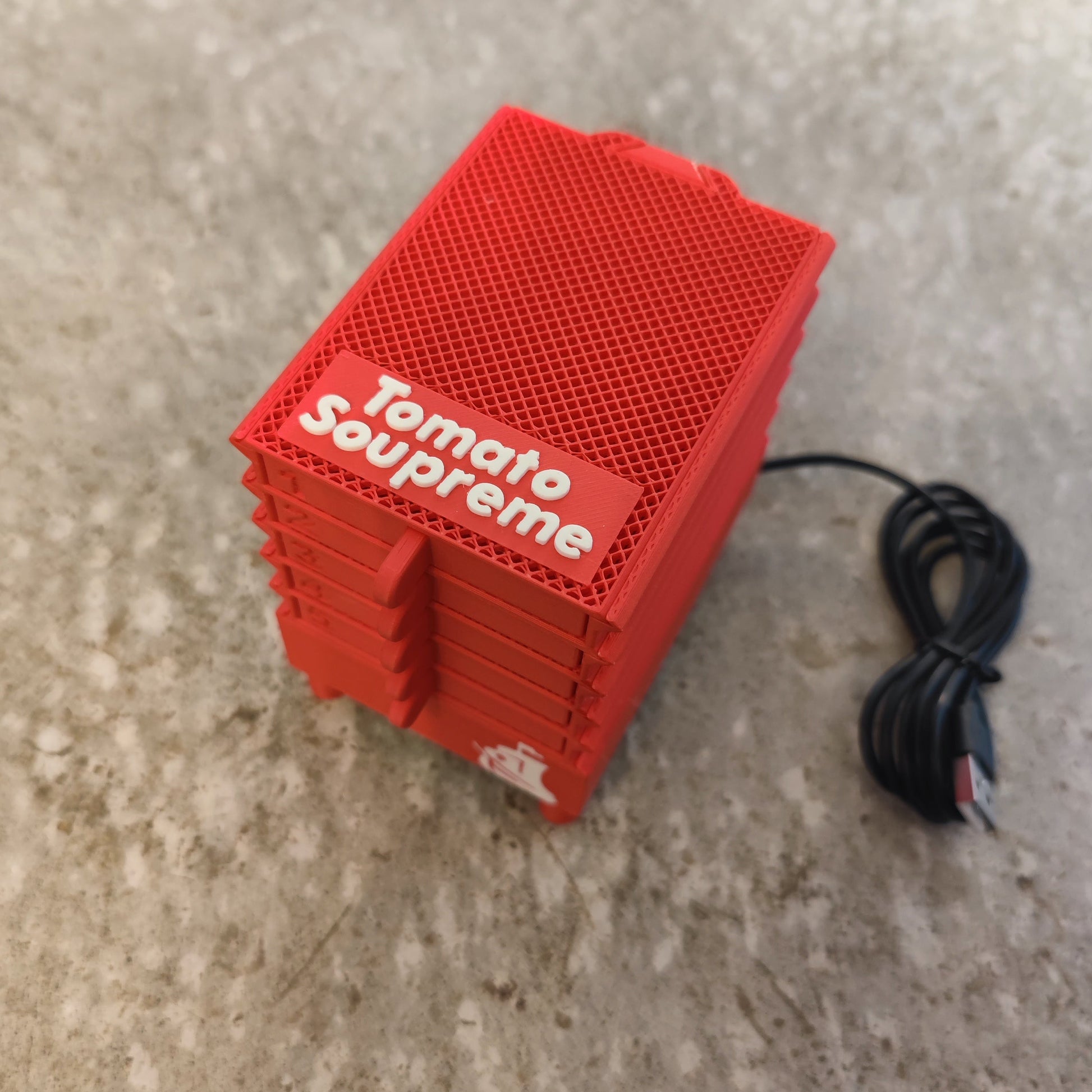 A compact red Seed Dryer - Tomato Soupreme from PepperMerchant.net rests on a gray surface with a black USB cable, highlighting innovative technology for efficient, gentle seed preservation.