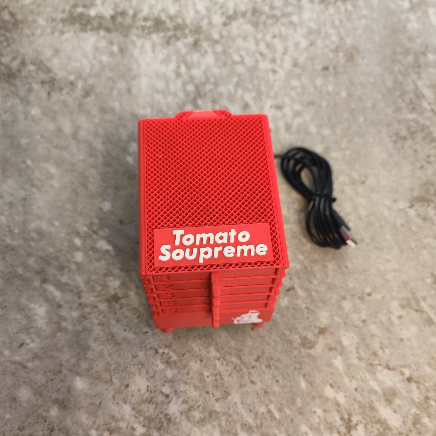 The PepperMerchant.net Seed Dryer - Tomato Soupreme, a compact red plastic crate with a textured lid, sits beside a coiled black USB cable on a light surface, suggesting advanced seed saving technology.
