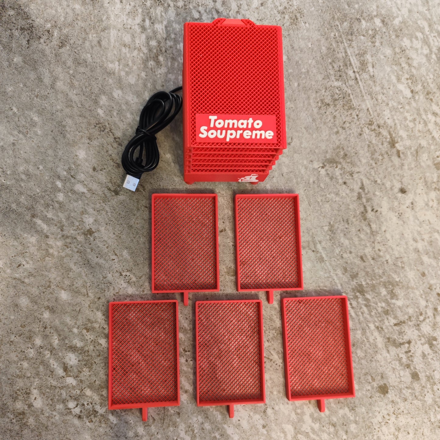 A red rectangular PepperMerchant.net Seed Dryer - Tomato Soupreme with a USB cable and five mesh trays are displayed on a textured gray surface, highlighting this innovative USB powered seed dryer.