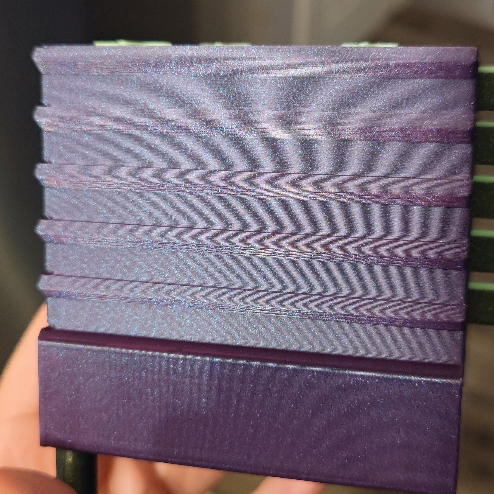 Close-up of the Seed Dryer - Nightshade (Glows) by PepperMerchant.net, a purple, rectangular, ridged device with a shimmery surface and stacked horizontal layers for advanced heatless seed drying, held against a blurred green background.