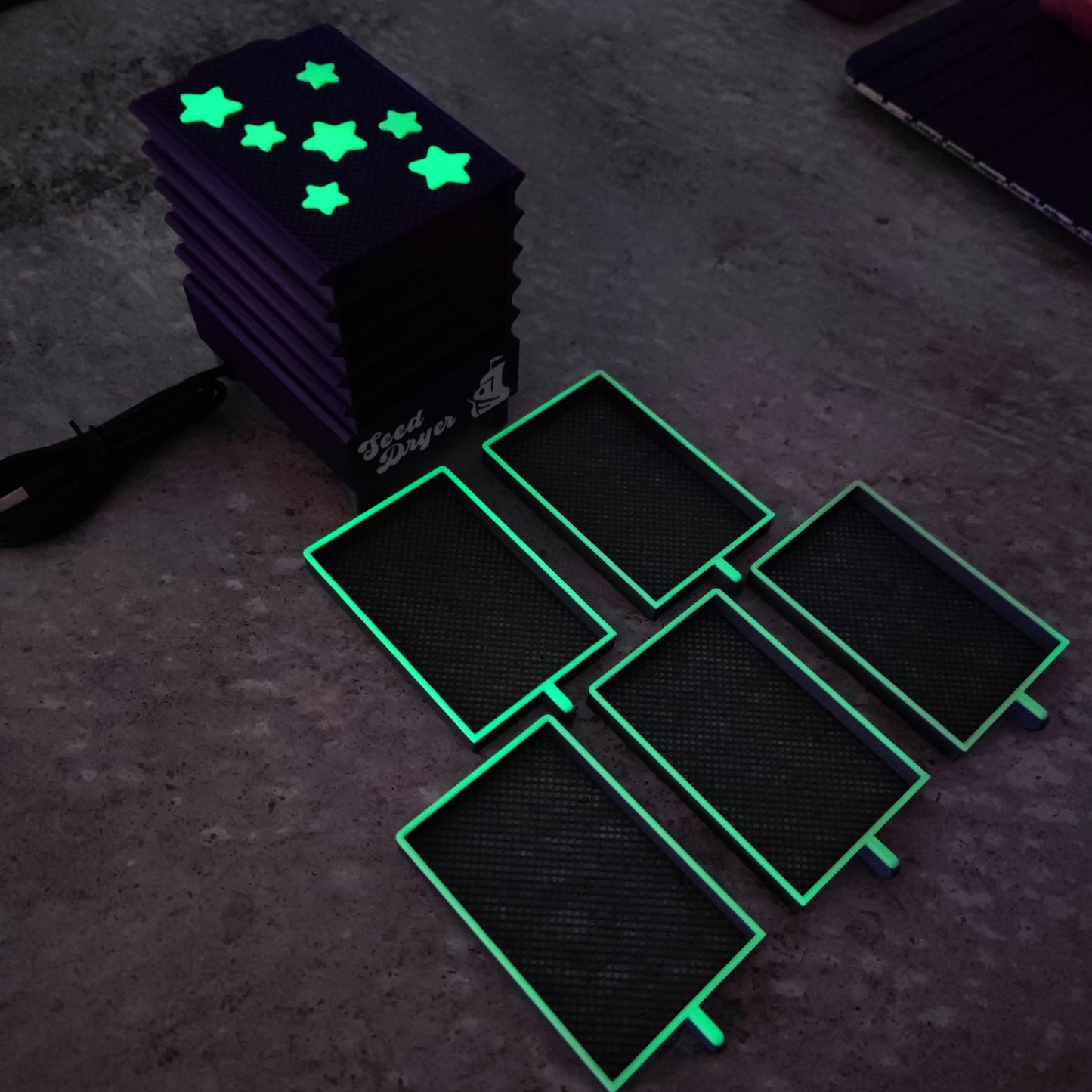 Five PepperMerchant.net Seed Dryer - Nightshade (Glows) covers with bright green edges sit beside a stack of star-cutout glow-in-the-dark covers. On the left, a USB-powered seed dryer cable is partially visible.