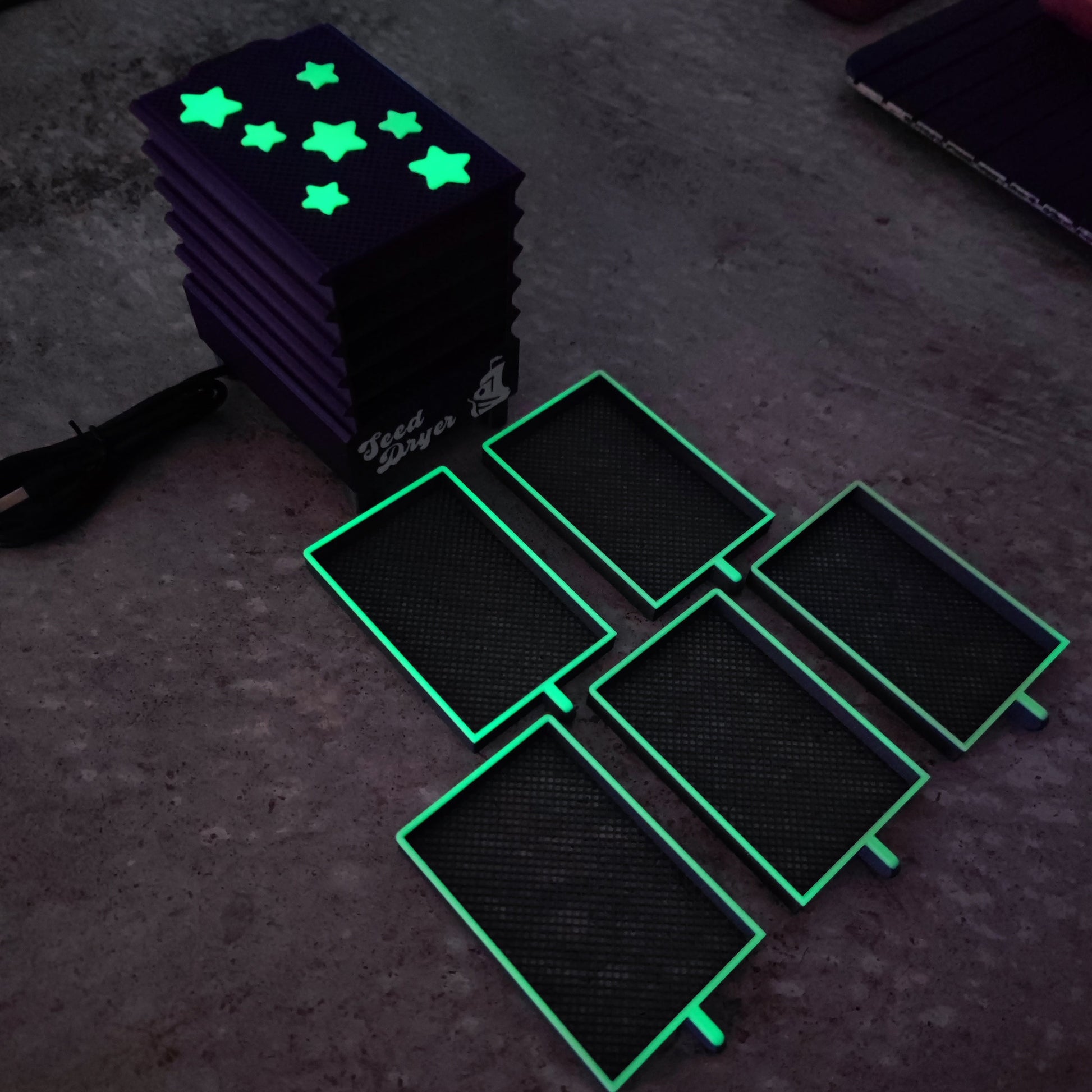 Five PepperMerchant.net Seed Dryer - Nightshade (Glows) covers with bright green edges sit beside a stack of star-cutout glow-in-the-dark covers. On the left, a USB-powered seed dryer cable is partially visible.