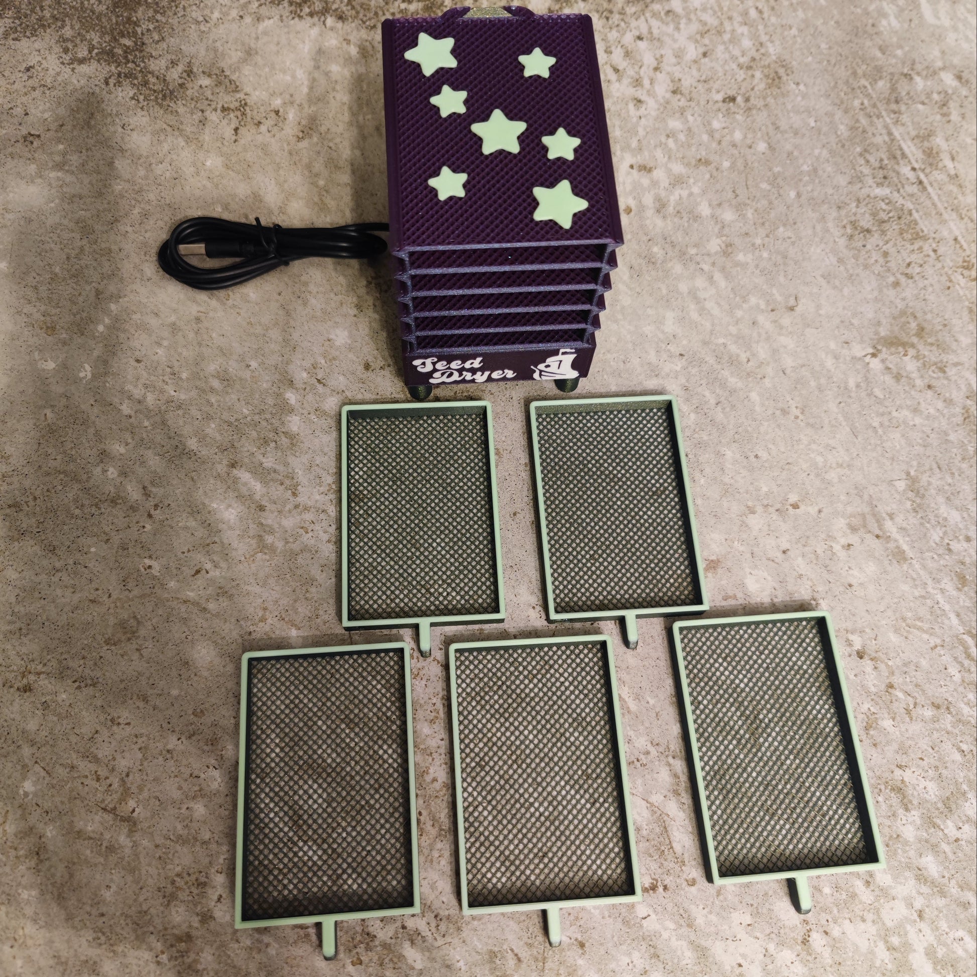 The Seed Dryer - Nightshade (Glows) by PepperMerchant.net features a purple design with star-shaped top vents, a black power cord, and five mesh trays—an innovative seed saving solution shown on a textured gray surface.