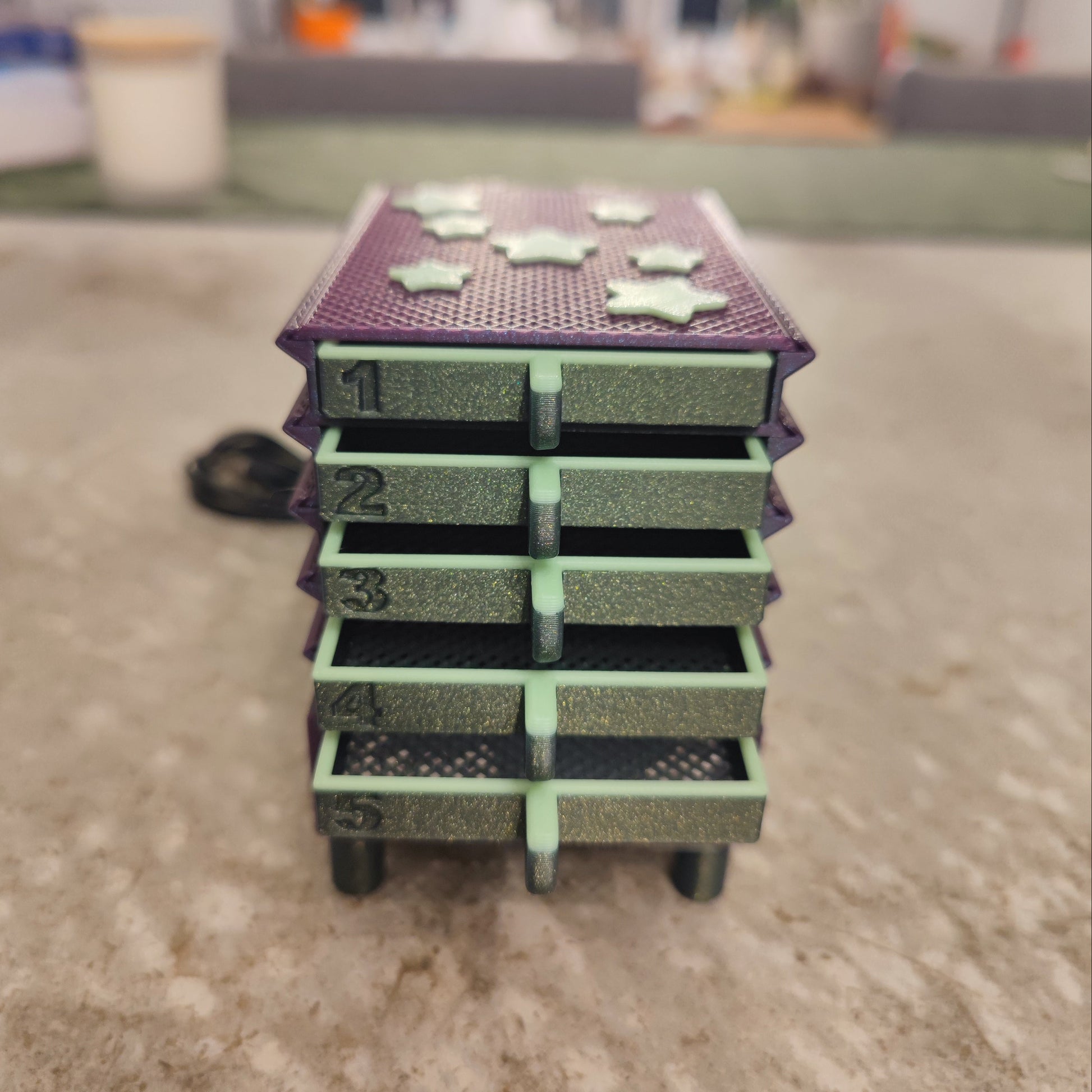 A close-up of a five-drawer purple organizer with green drawers and star accents sits on a gray surface—ideal for storing accessories for the PepperMerchant.net Seed Dryer - Nightshade (Glows).