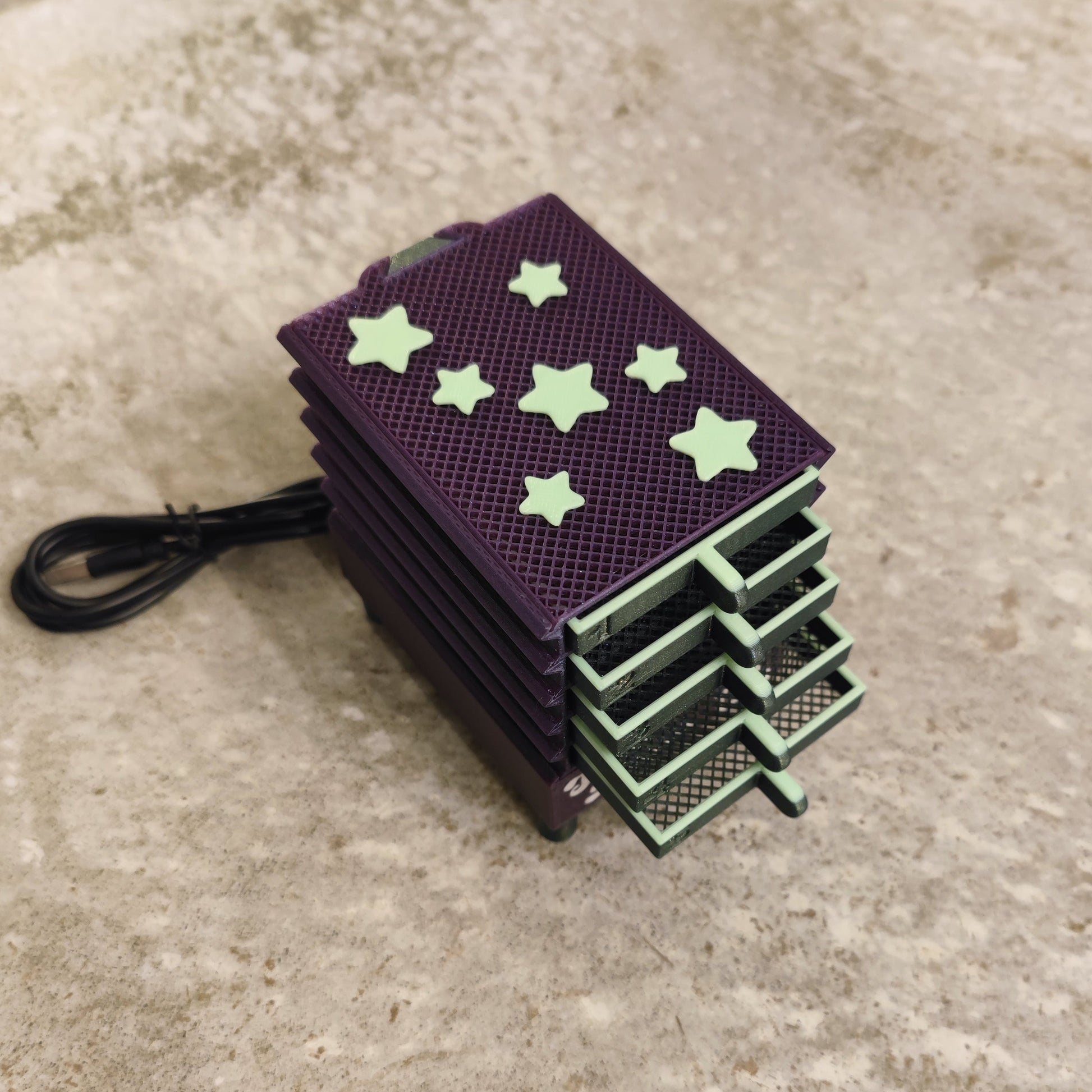 The Seed Dryer - Nightshade (Glows) by PepperMerchant.net is a small, purple rectangular USB-powered device with green star-shaped accents and a stack-like design, connected by a black cable on a textured beige surface.