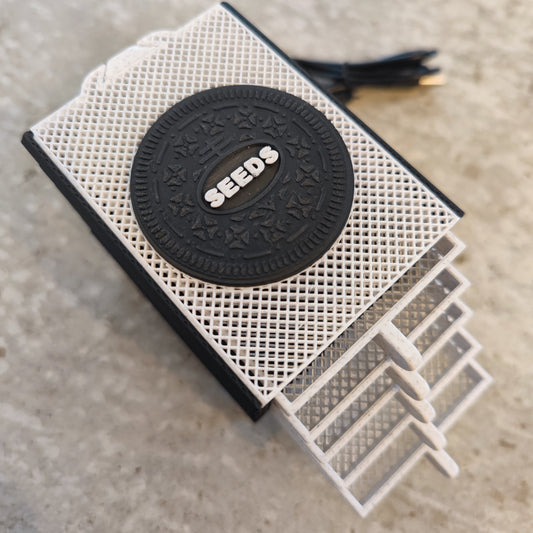 The Seed Dryer - Cookies & Cream by PepperMerchant.net is a 3D-printed Oreo-style device with "SEEDS" in the center, four sliding trays for seeds, and a USB-powered black cord, shown resting on a speckled surface.