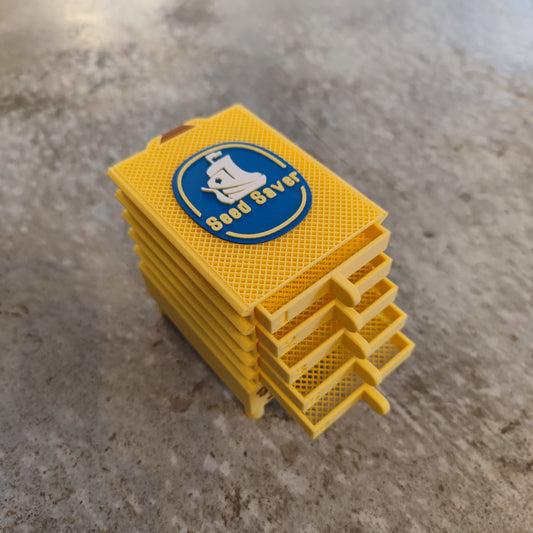 The Seed Dryer - Banana by PepperMerchant.net is a stack of yellow, rectangular trays with mesh bottoms and "Seed Saver" printed on top, designed for innovative seed drying, shown on a speckled gray surface.