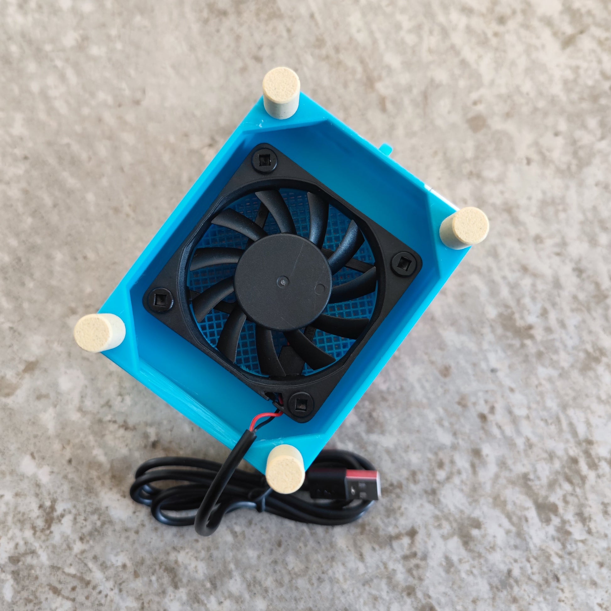 A small black cooling fan, housed in a blue 3D-printed case with four beige feet and a black power cable, sits on a gray textured surface, showcasing the Seed Dryer - Rocket Pop by PepperMerchant.net—an innovative and essential USB-powered seed drying device for seed saving.