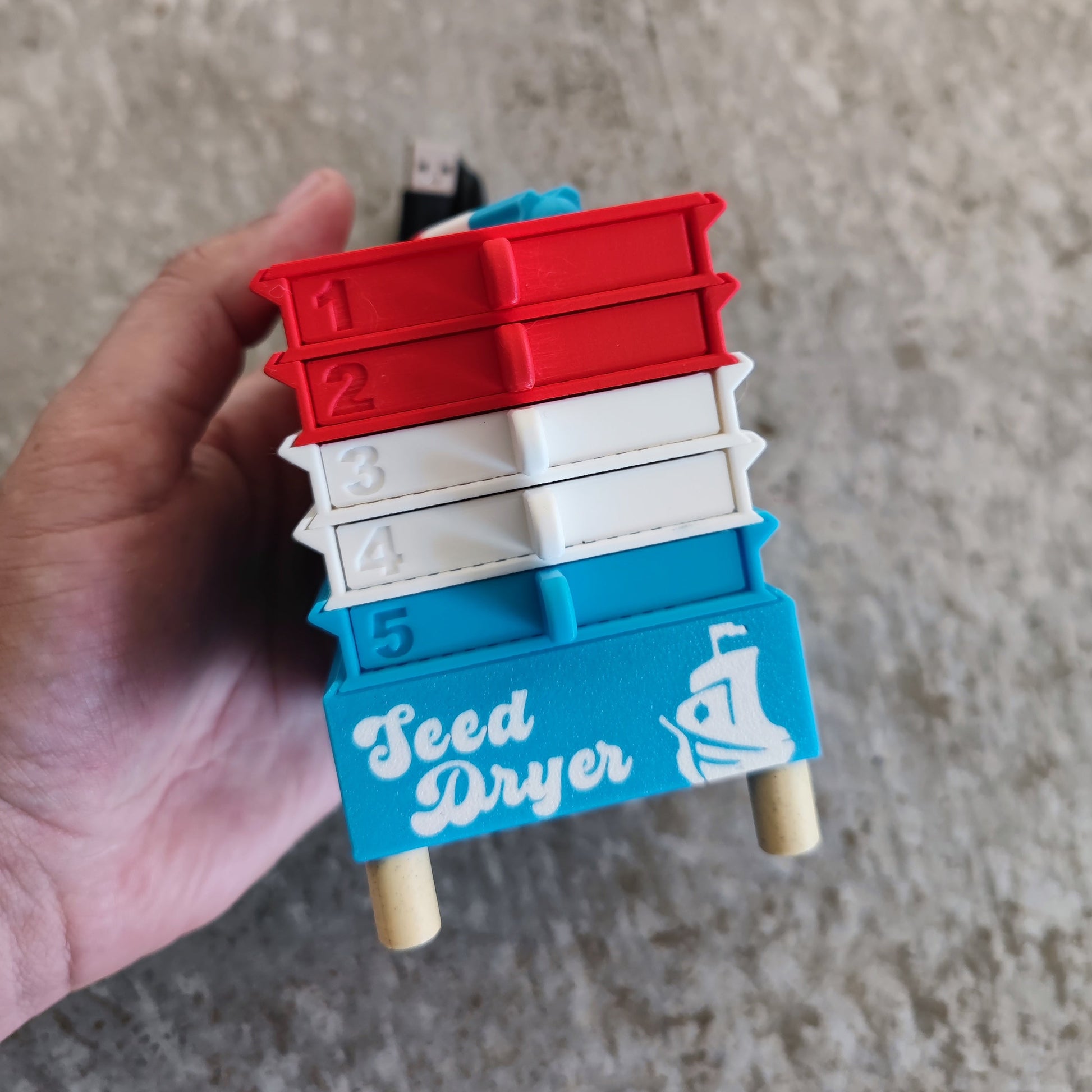 A hand holds a colorful device labeled "Seed Dryer - Rocket Pop" from PepperMerchant.net, featuring five numbered drawers in red, white, and blue with a fan icon—representing a USB-powered seed dryer using advanced seed saving technology. A USB cable is visible at the top.