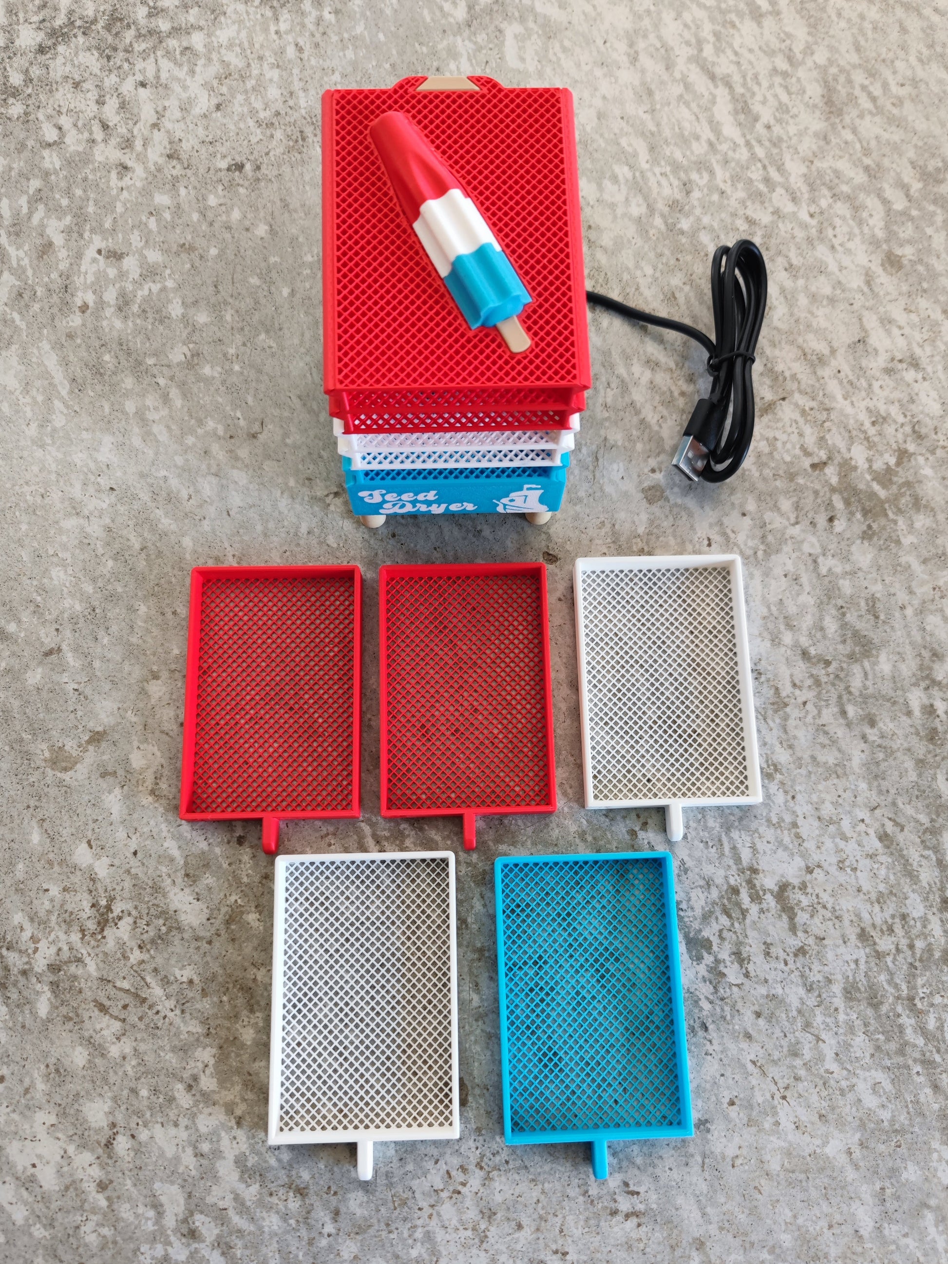 Showcasing innovative seed-saving technology, the Seed Dryer - Rocket Pop by PepperMerchant.net—a USB-powered mini appliance featuring a popsicle-shaped top—sits on a textured concrete surface. In front, four mesh trays in red, white, and blue highlight its compact and efficient design for drying seeds.