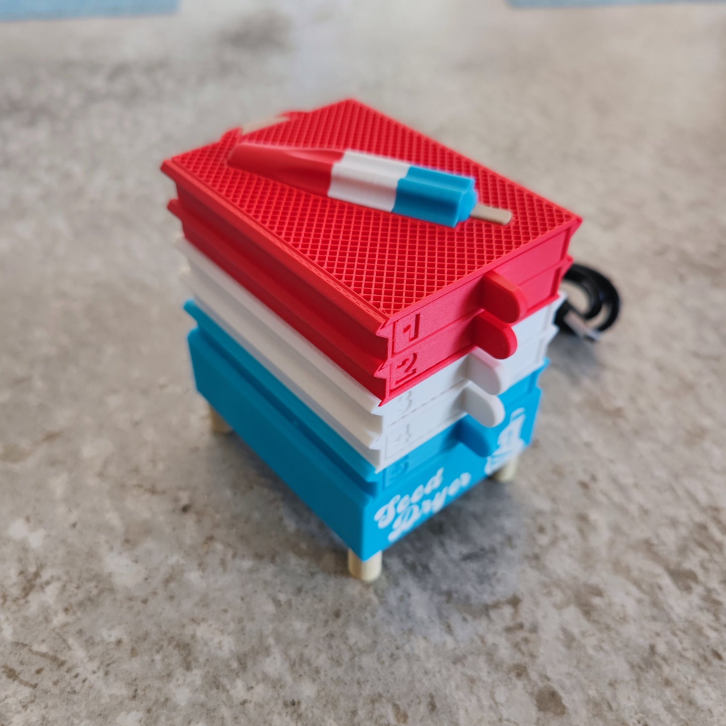 A small model of stacked red, white, and blue picnic coolers with a miniature red, white, and blue popsicle on top sits on a gray surface—echoing how the Seed Dryer - Rocket Pop from PepperMerchant.net delivers both efficiency and innovation to everyday seed saving.