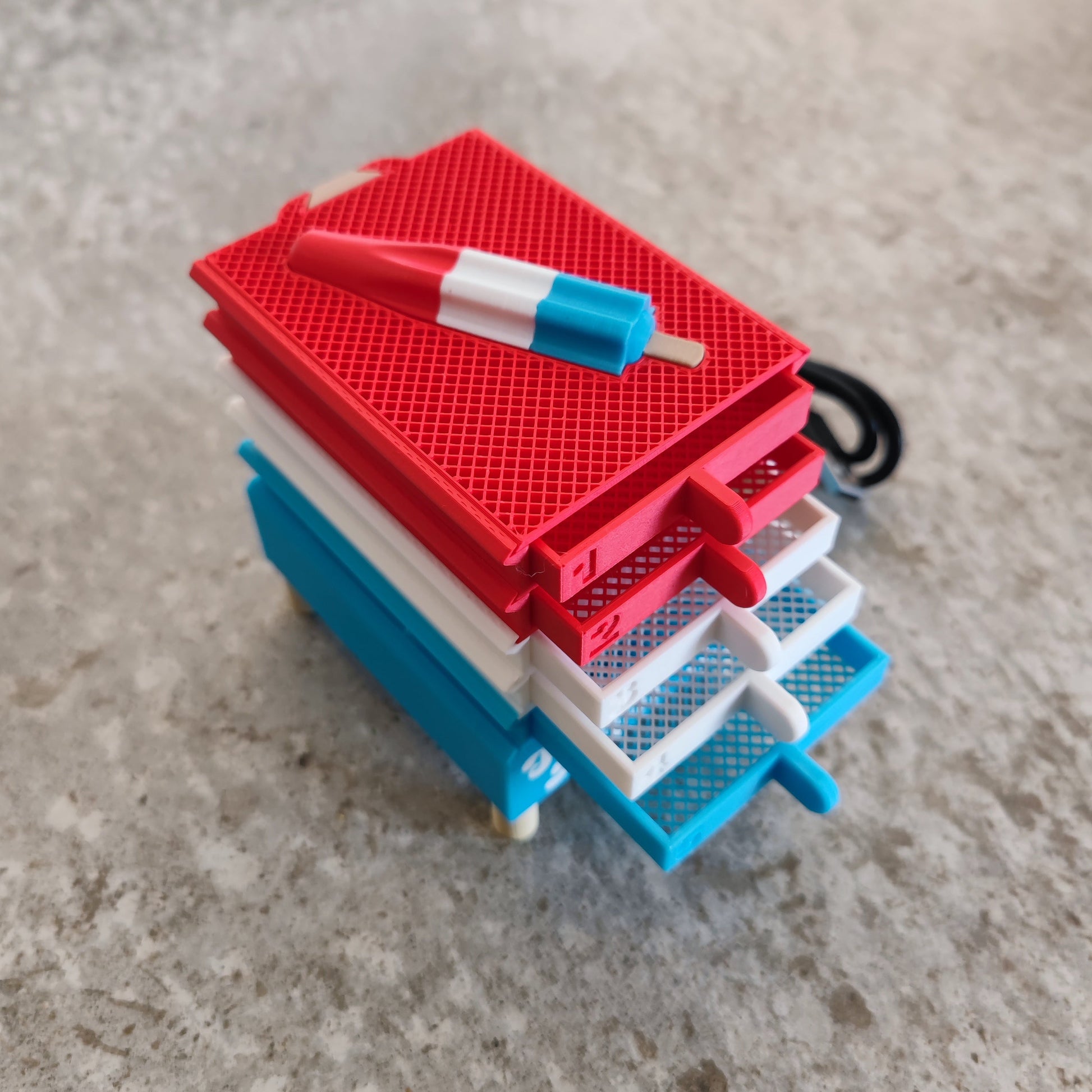 A Seed Dryer - Rocket Pop from PepperMerchant.net, featuring a stack of mini table tennis paddles in red, white, and blue with a matching mini paddle-shaped keychain on top, evokes the same compact efficiency found in modern seed saving technology—all neatly arranged on a gray textured surface.