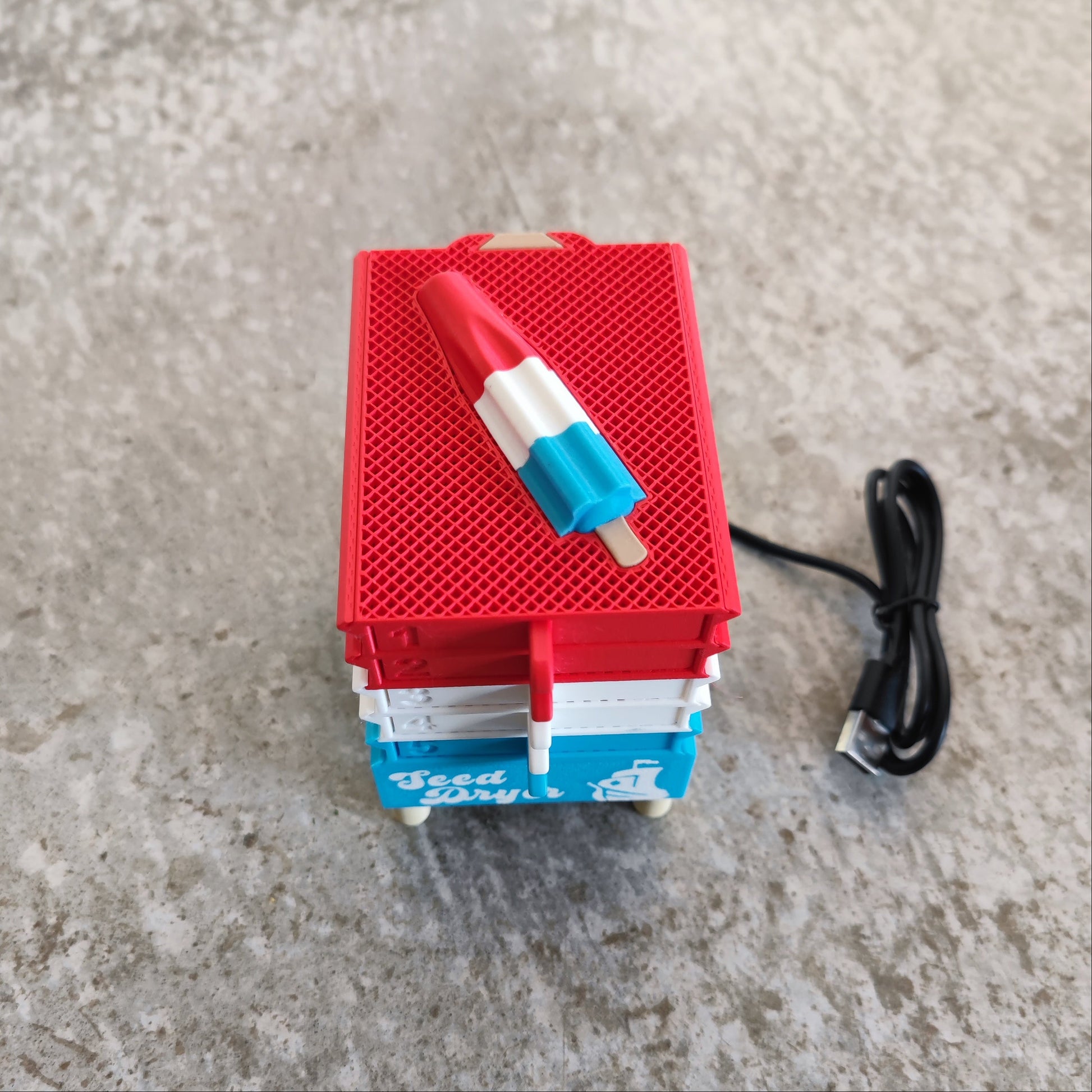 A USB cable connects to the Seed Dryer - Rocket Pop by PepperMerchant.net, a compact device inspired by modern seed-saving technology that resembles a stack of coolers with a red, white, and blue popsicle on top. It sits atop a textured gray surface.
