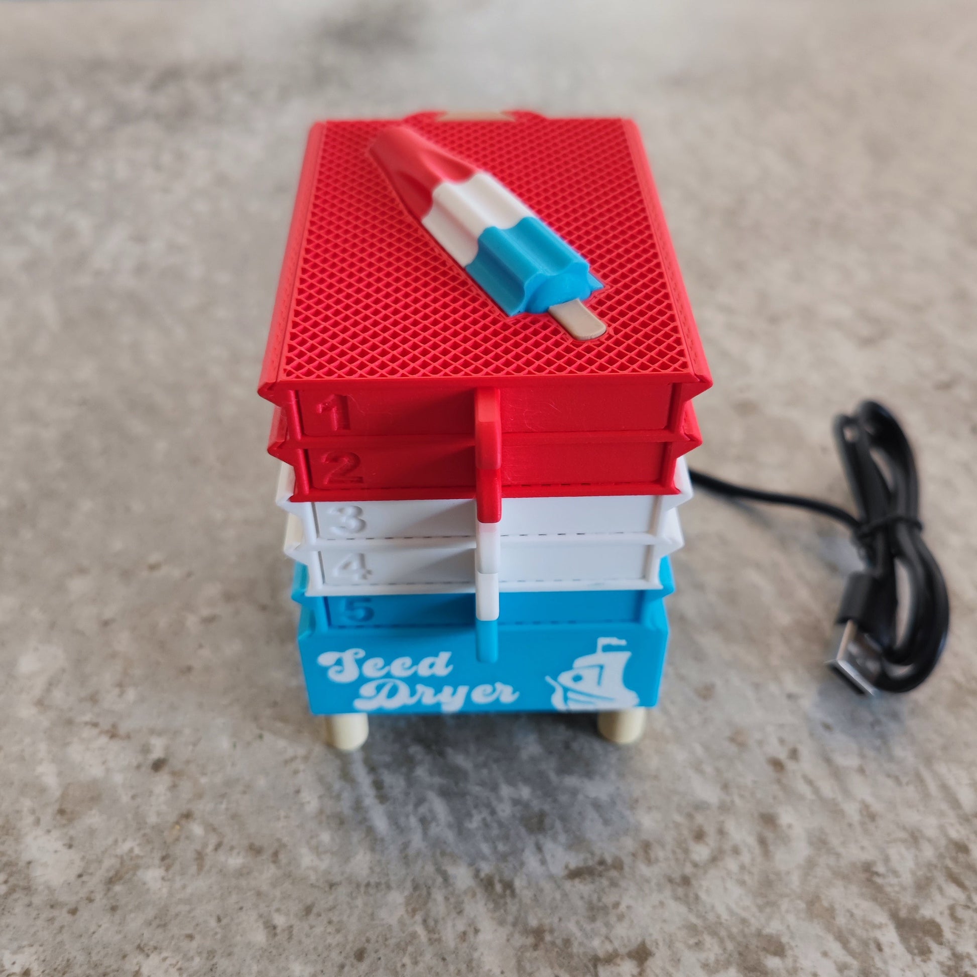 A small, colorful USB-powered seed dryer labeled "Seed Dryer – Rocket Pop" from PepperMerchant.net, featuring five numbered trays and a rocket pop–shaped object on top to highlight its innovative seed-saving technology, displayed on a gray surface.