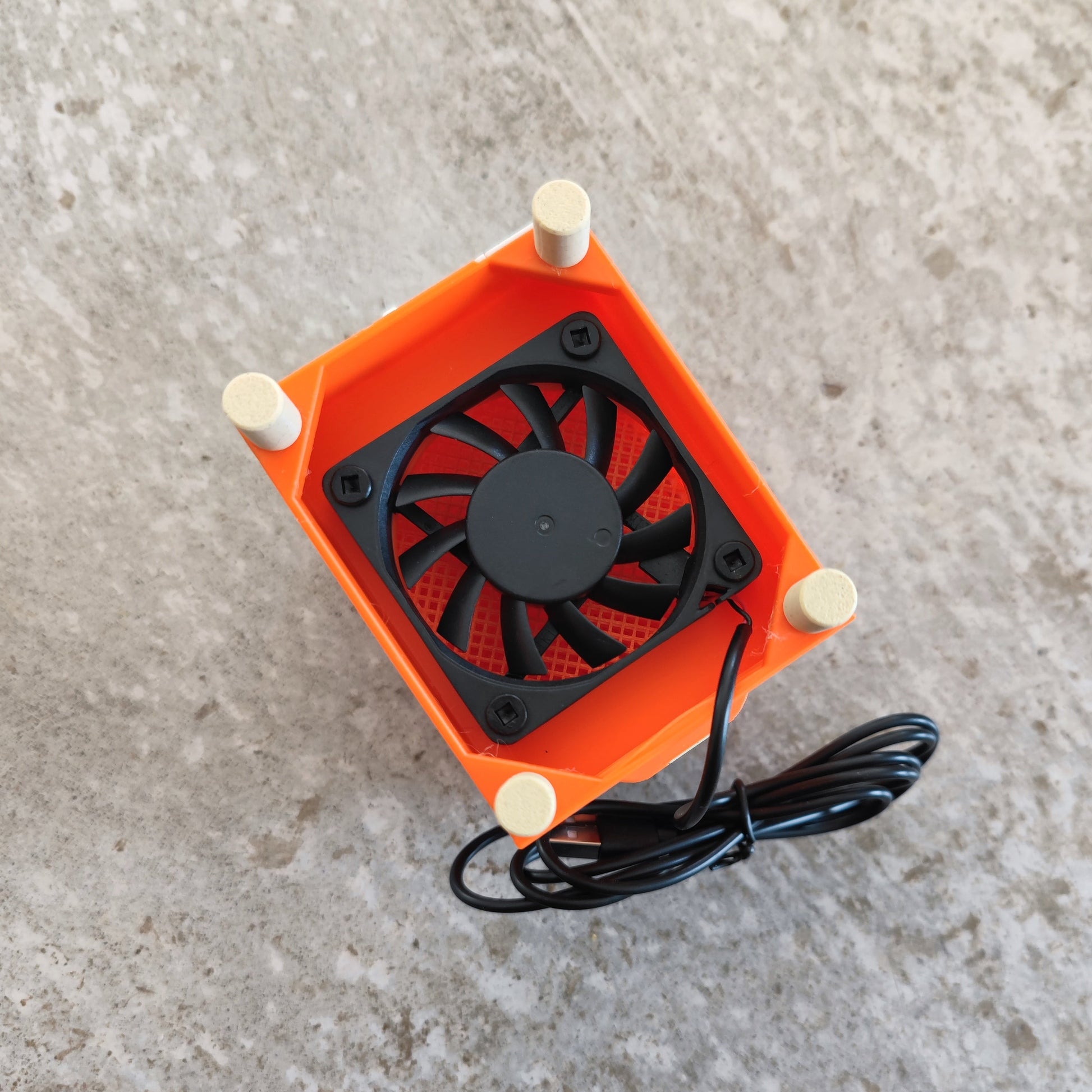 A compact black cooling fan encased in an orange plastic frame with four round feet and a black cable, branded as the "Seed Dryer - Creamsicle" by PepperMerchant.net, sits on a textured gray surface—an innovative USB-powered seed dryer that is an essential tool for modern seed saving technology.