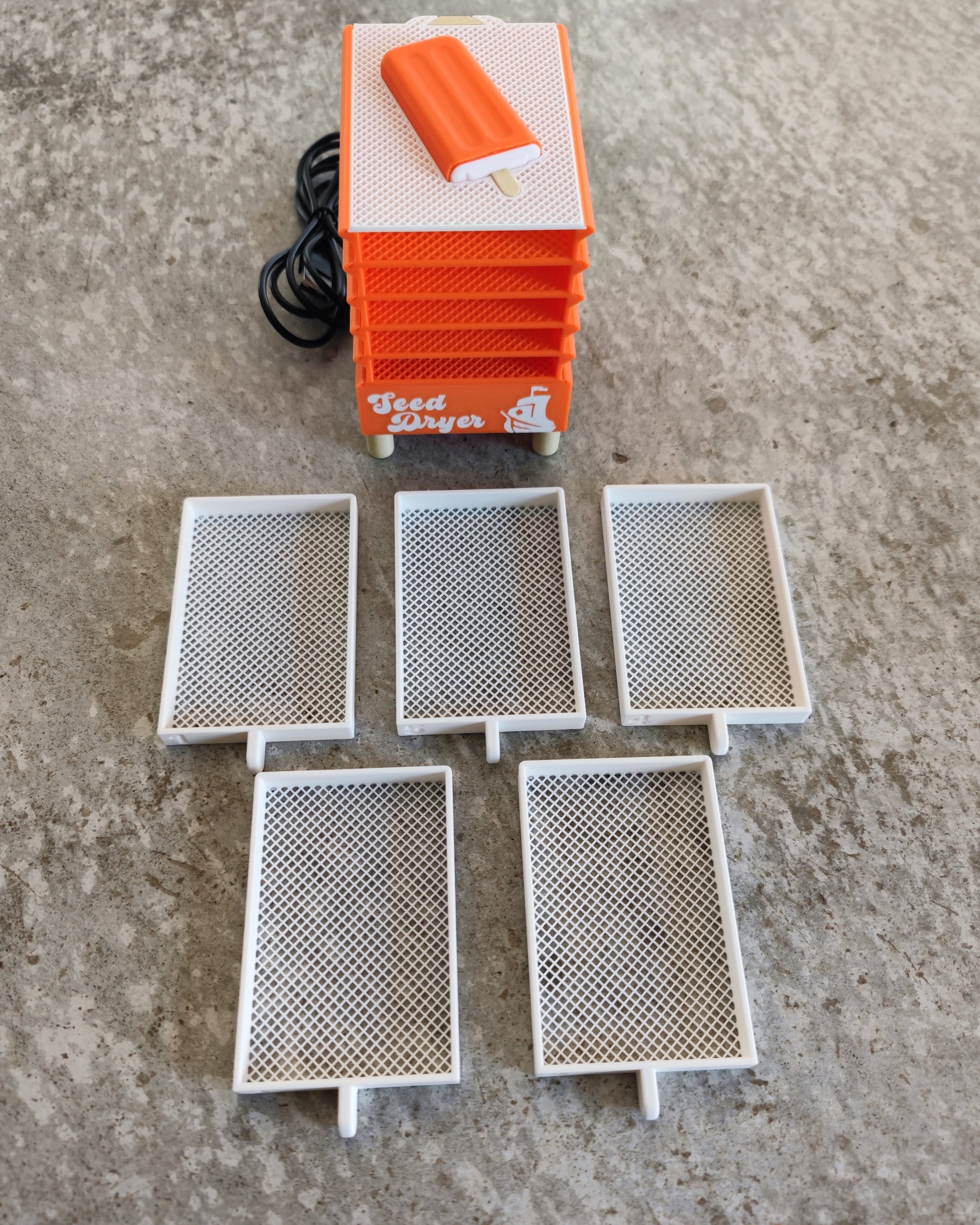 A Seed Dryer - Creamsicle by PepperMerchant.net with a cord is displayed on a gray surface, accompanied by five removable white mesh trays arranged in front, showcasing modern seed saving technology.