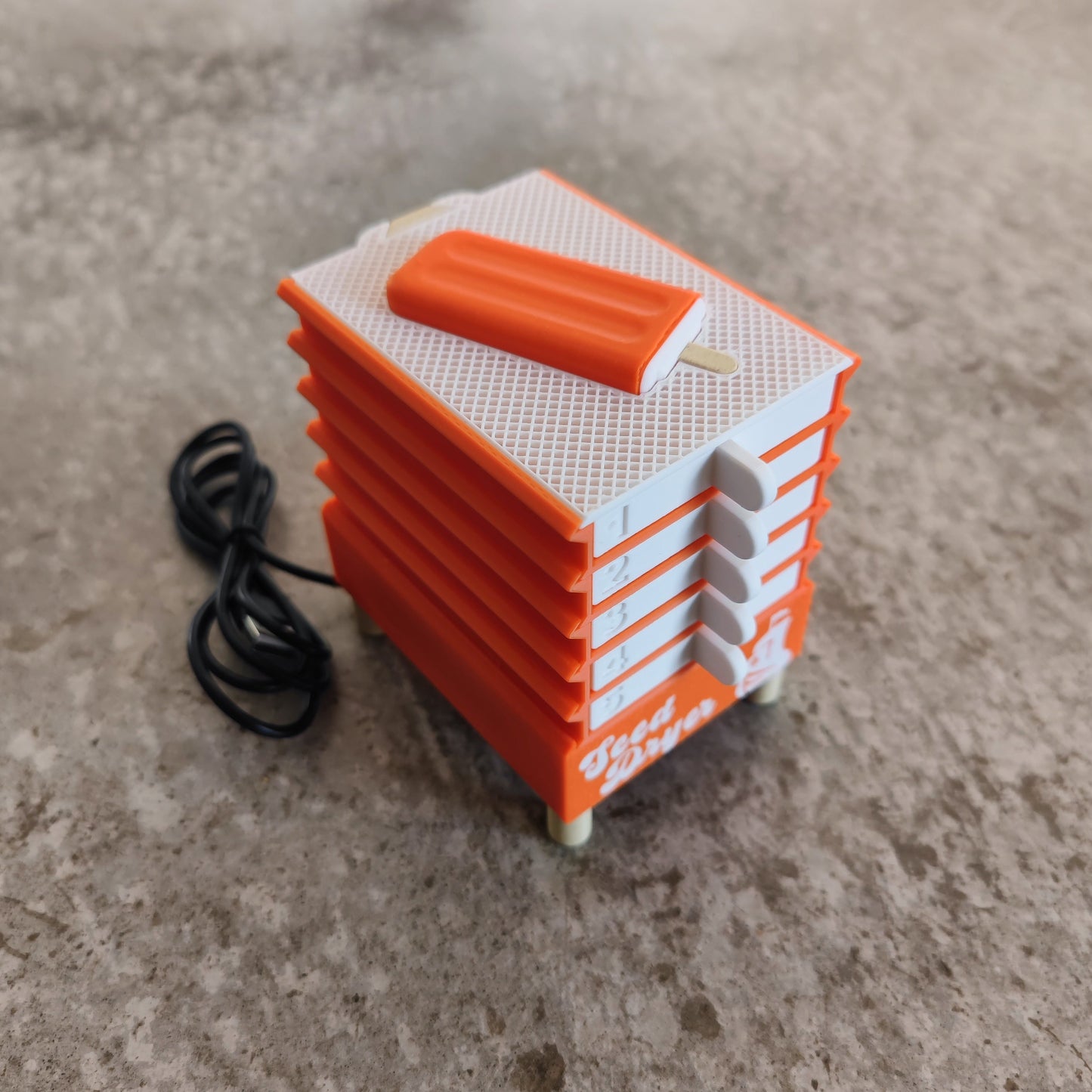 A PepperMerchant.net Seed Dryer - Creamsicle, a small orange and white USB-powered device resembling stacked popsicles with a cord attached, sits on a gray textured surface.