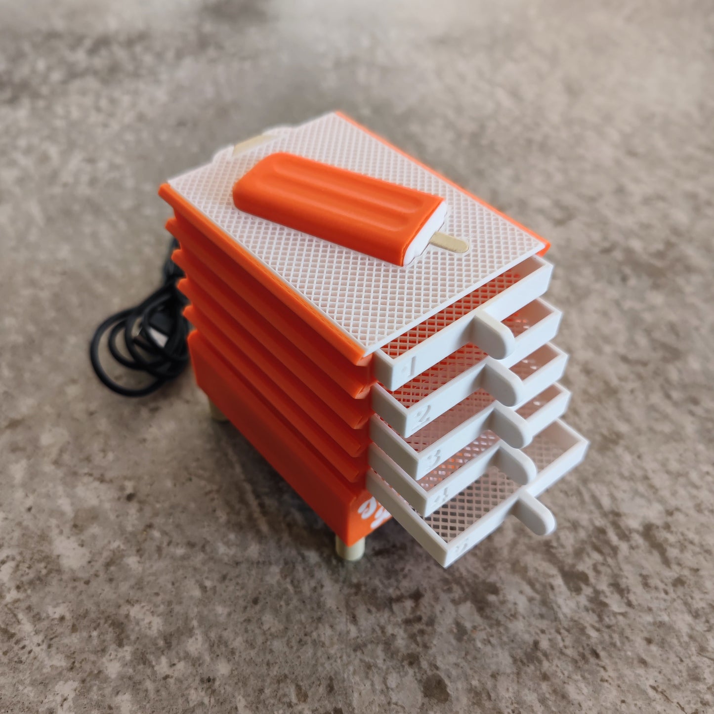 A miniature orange and white plastic popsicle-shaped object with five stacked trays and a black cord sits on a textured surface—actually, this is the Seed Dryer - Creamsicle by PepperMerchant.net, a clever USB-powered seed dryer designed for modern seed saving technology.