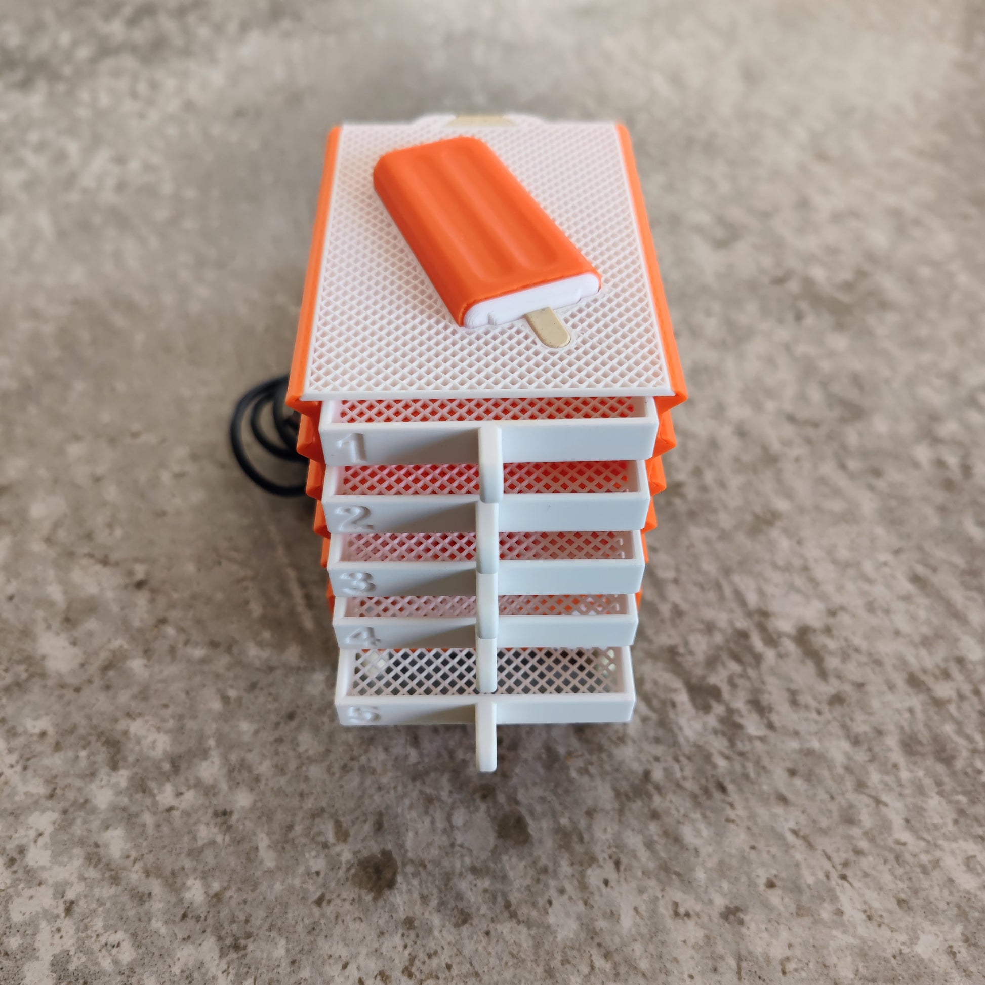A stack of five orange and white mesh trays, part of the PepperMerchant.net Seed Dryer - Creamsicle—an innovative USB-powered seed dryer—sits atop a speckled gray surface with a small orange popsicle-shaped object on top.