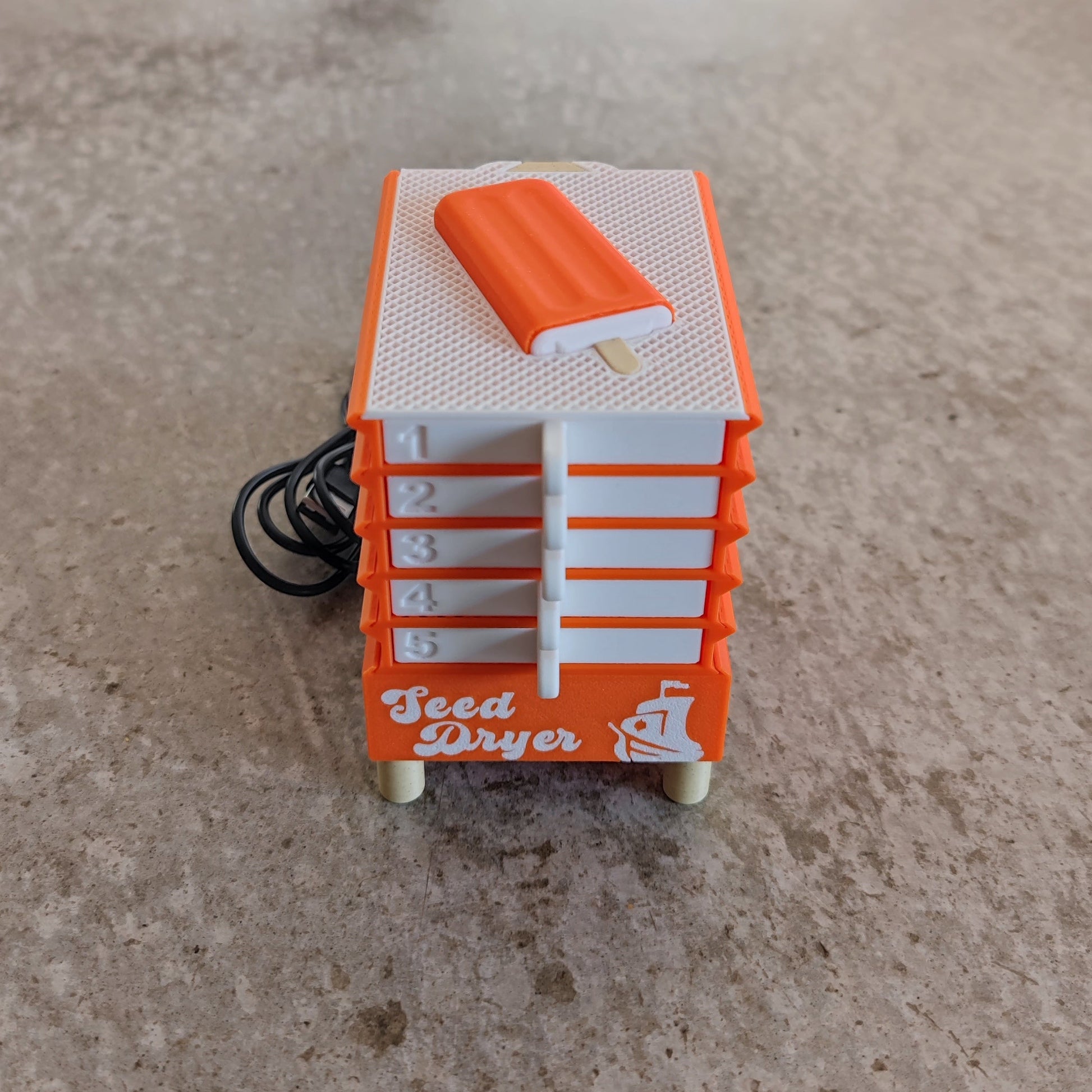 A PepperMerchant.net Seed Dryer - Creamsicle in small orange and white, featuring five numbered drawers, a white knob, and a black power cord, sits on a beige surface.