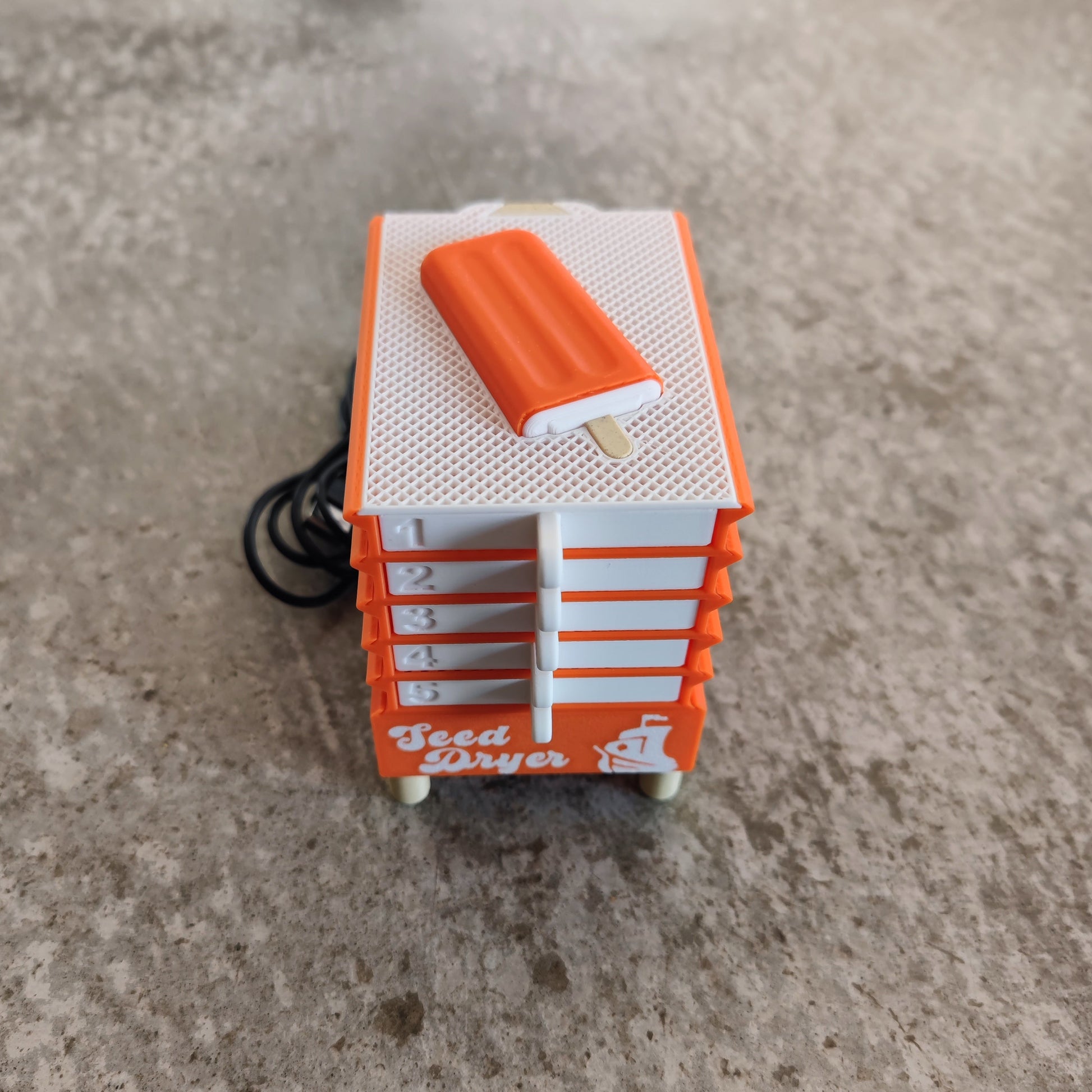 A compact orange and white device labeled "Seed Dryer - Creamsicle" from PepperMerchant.net, featuring numbered drawers and a popsicle-shaped accent on top, sits on a gray surface and exemplifies innovative seed-saving technology. This USB-powered seed dryer provides efficient and convenient drying for preserving seeds.