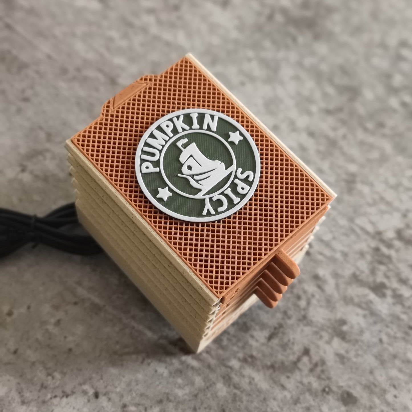 The Seed Dryer - Pumpkin Spicy *Limited Edition* from PepperMerchant.net, featuring tan sides, orange grid vents, a pumpkin spice logo, and a black cable, rests on a gray stone surface—perfect for the dedicated seed saver.