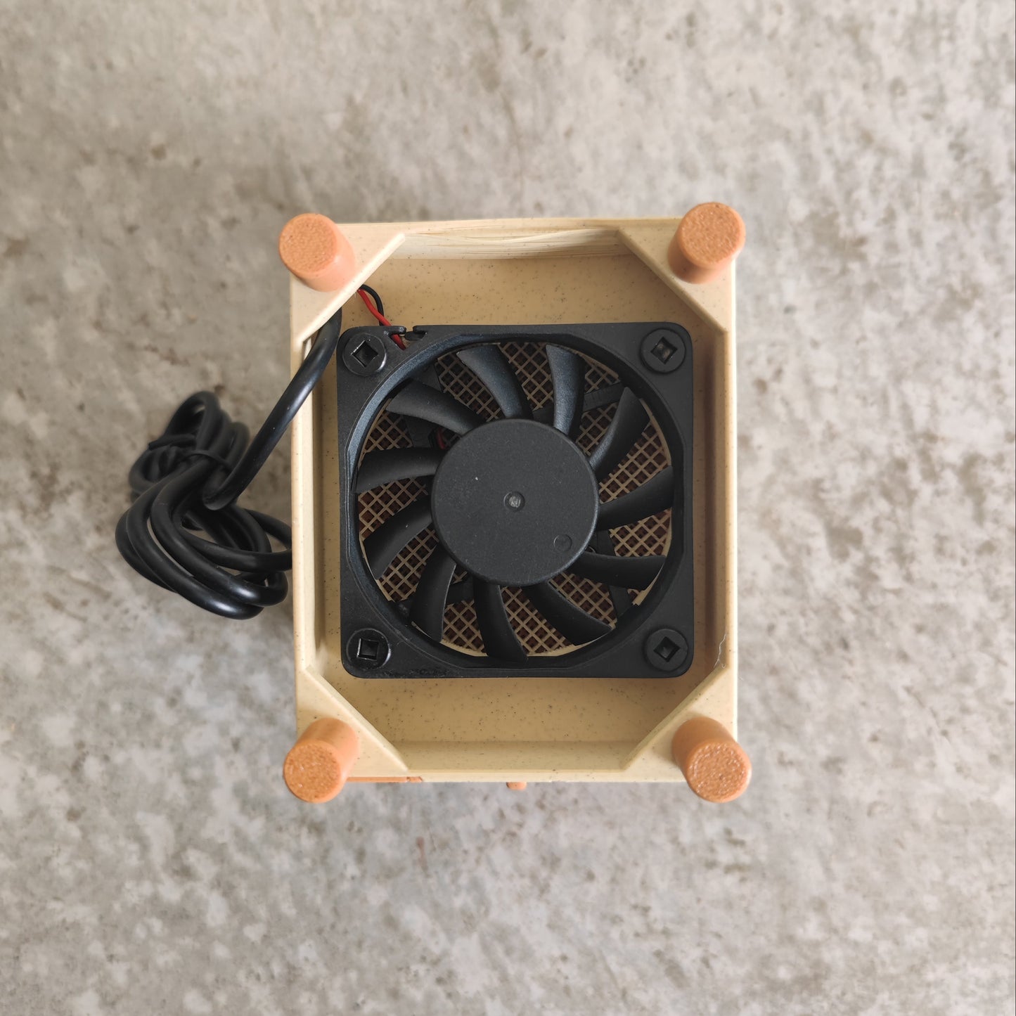 A compact black cooling fan with a black cord is mounted inside a beige rectangular frame accented by four orange corner caps, resting on a gray textured surface—making the PepperMerchant.net Seed Dryer - Pumpkin Spicy *Limited Edition* the perfect USB-powered choice for any dedicated seed saver.