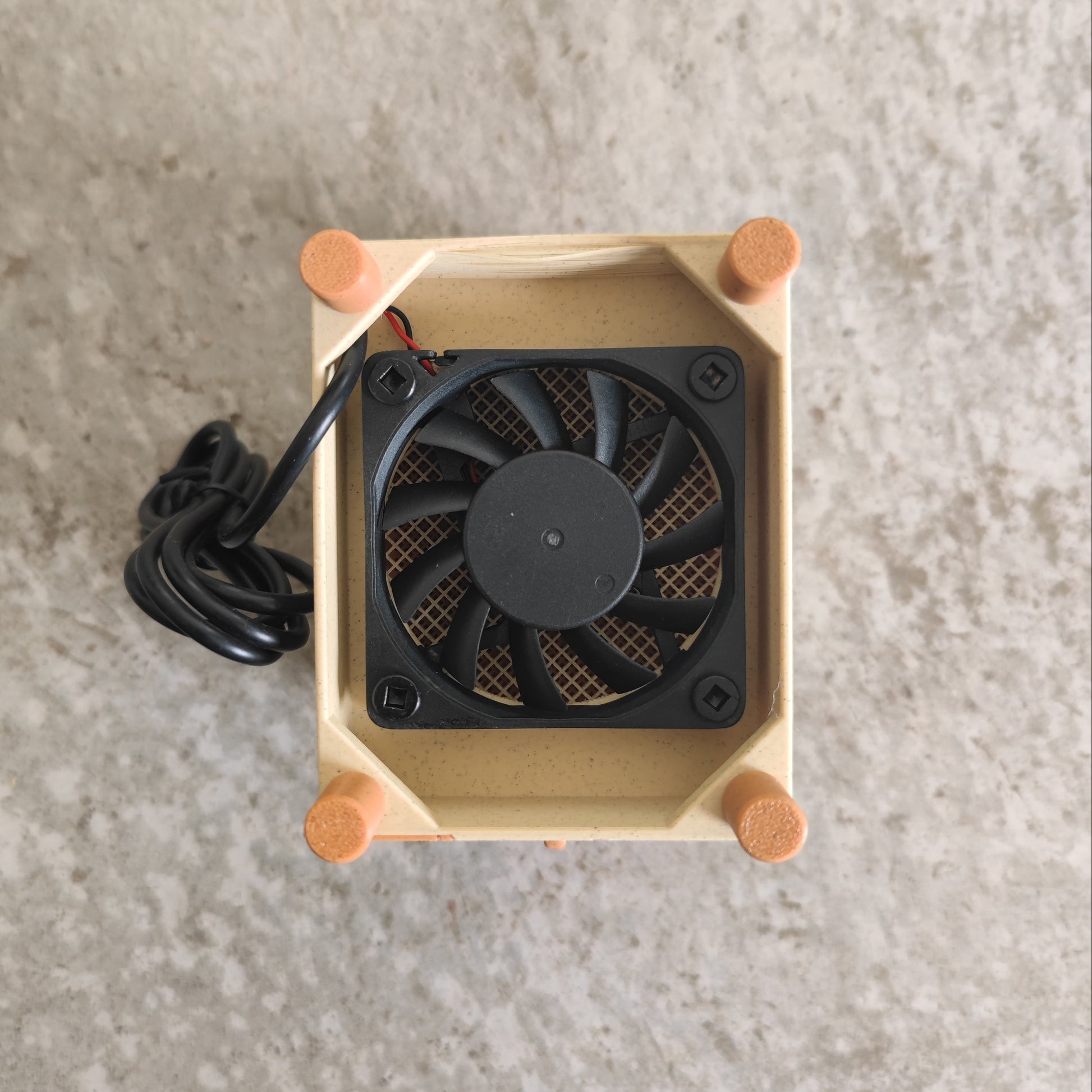 A compact black cooling fan with a black cord is mounted inside a beige rectangular frame accented by four orange corner caps, resting on a gray textured surface—making the PepperMerchant.net Seed Dryer - Pumpkin Spicy *Limited Edition* the perfect USB-powered choice for any dedicated seed saver.