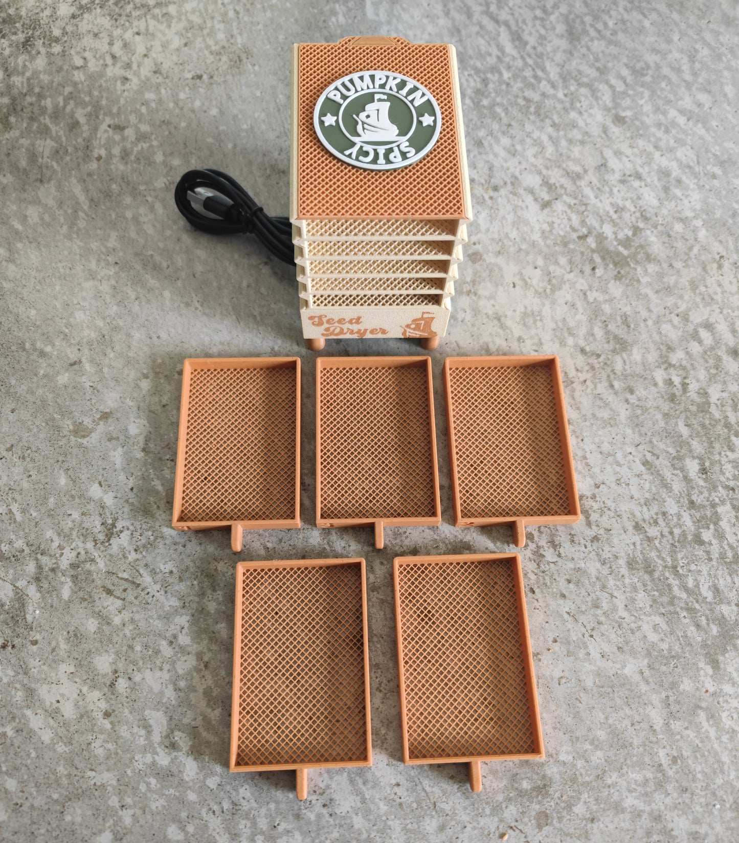Five orange honeycomb-patterned trays are arranged on a concrete surface in front of a white and orange stacked device labeled “Seed Dryer - Pumpkin Spicy *Limited Edition*” from PepperMerchant.net, with a visible power cord, showcasing an innovative USB-powered seed dryer ideal for dedicated seed savers.