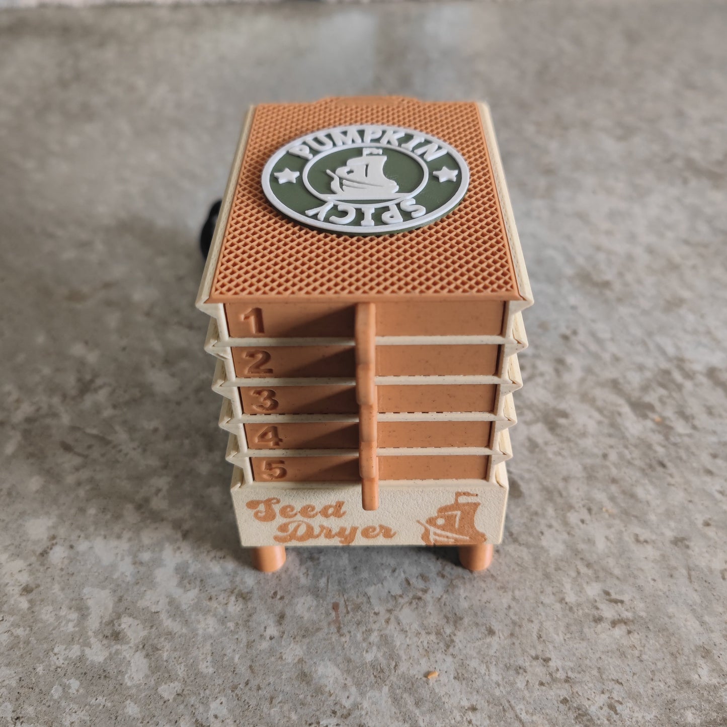 Here’s the revised sentence using your product data: The PepperMerchant.net Seed Dryer - Pumpkin Spicy *Limited Edition* is a compact, USB-powered seed dryer featuring five numbered trays and “Seed Dryer” displayed on the front. A circular “Pumpkin Spicy” logo decorates the top, emphasizing its distinctive pumpkin spice-themed design. Shown on a textured gray surface, this device is ideal for dedicated seed savers.