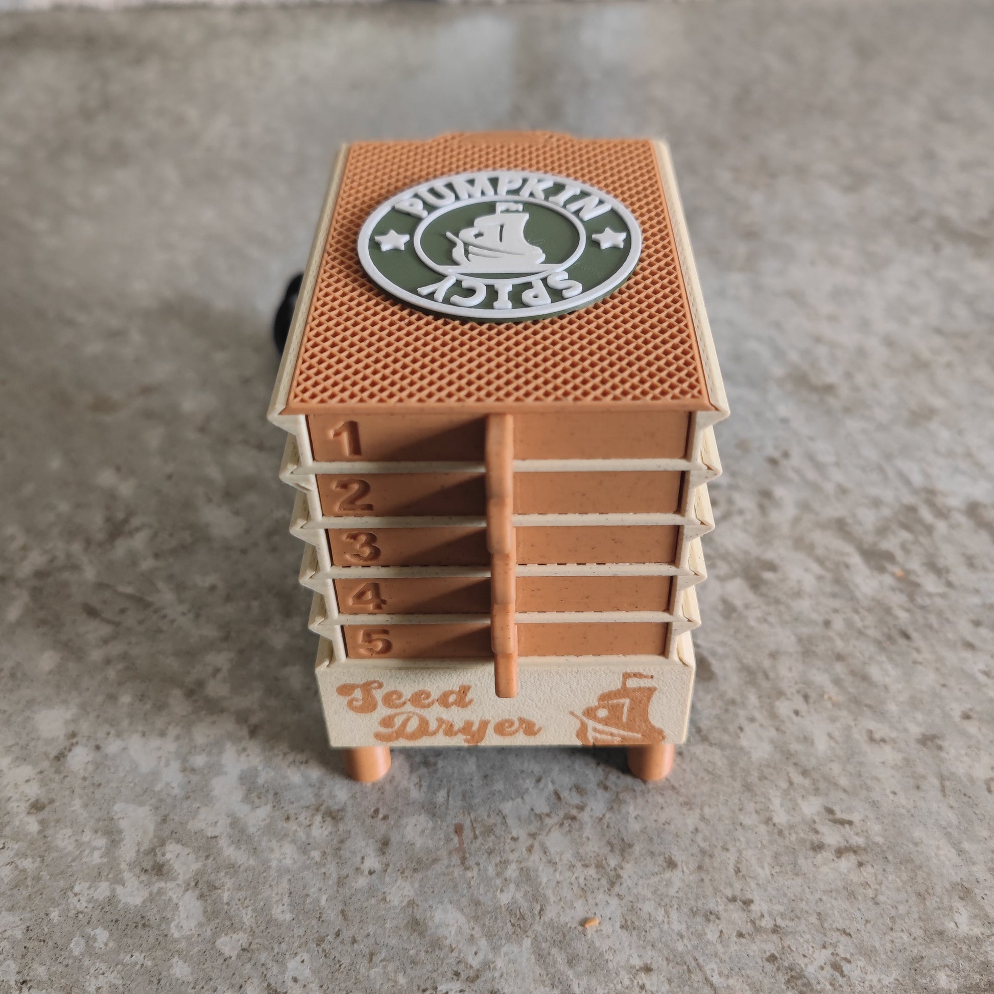 Here’s the revised sentence using your product data: The PepperMerchant.net Seed Dryer - Pumpkin Spicy *Limited Edition* is a compact, USB-powered seed dryer featuring five numbered trays and “Seed Dryer” displayed on the front. A circular “Pumpkin Spicy” logo decorates the top, emphasizing its distinctive pumpkin spice-themed design. Shown on a textured gray surface, this device is ideal for dedicated seed savers.