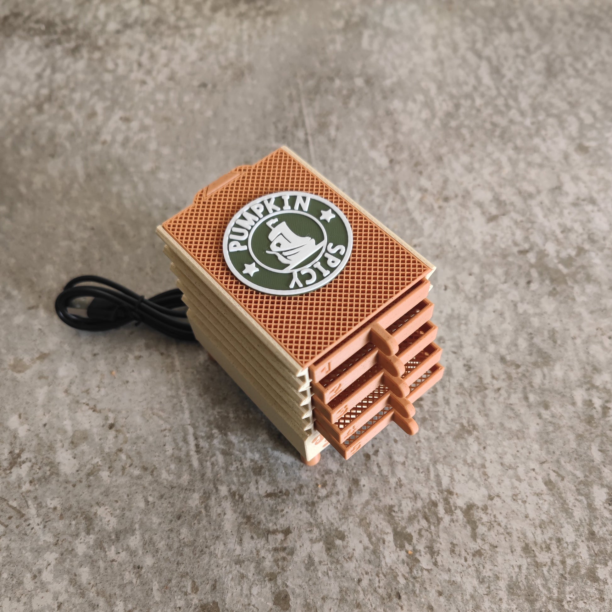 A tan and orange Seed Dryer—Pumpkin Spicy *Limited Edition* by PepperMerchant.net, featuring a vented design and distinctive “Pumpkin Spicy” branding, sits on a concrete surface. The device, reminiscent of an air freshener, has a black power cord attached to its side.