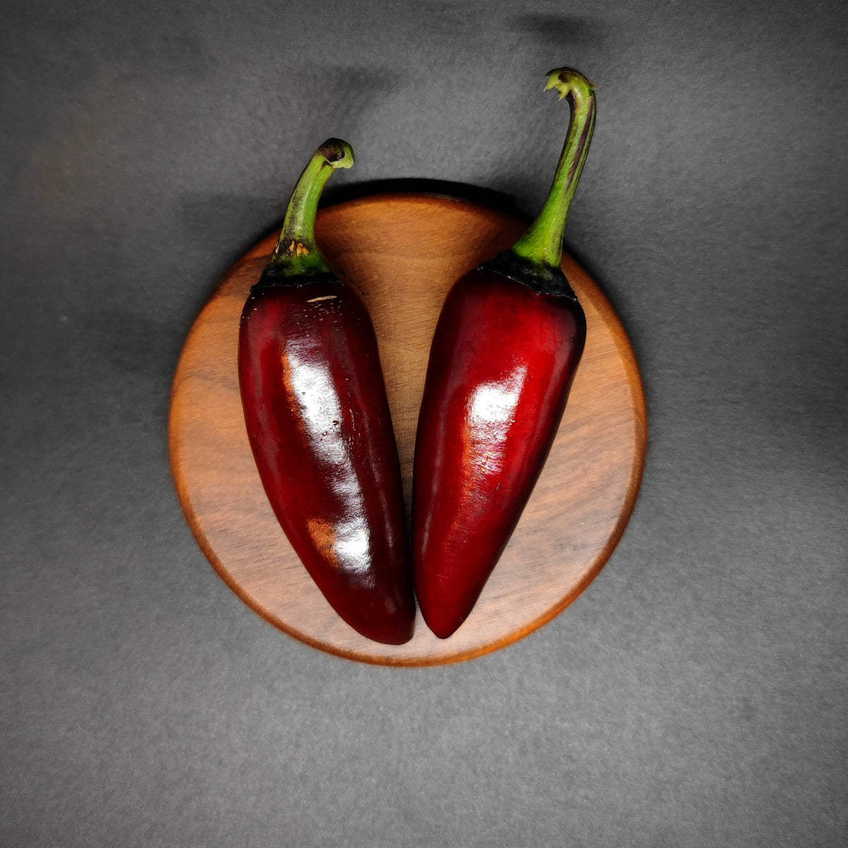 Two dark red Ruby Jalapeno peppers with green stems rest side by side on a round wooden plate, placed on a dark surface under dramatic lighting—an impressive showcase for unique seeds from PepperMerchant.net.