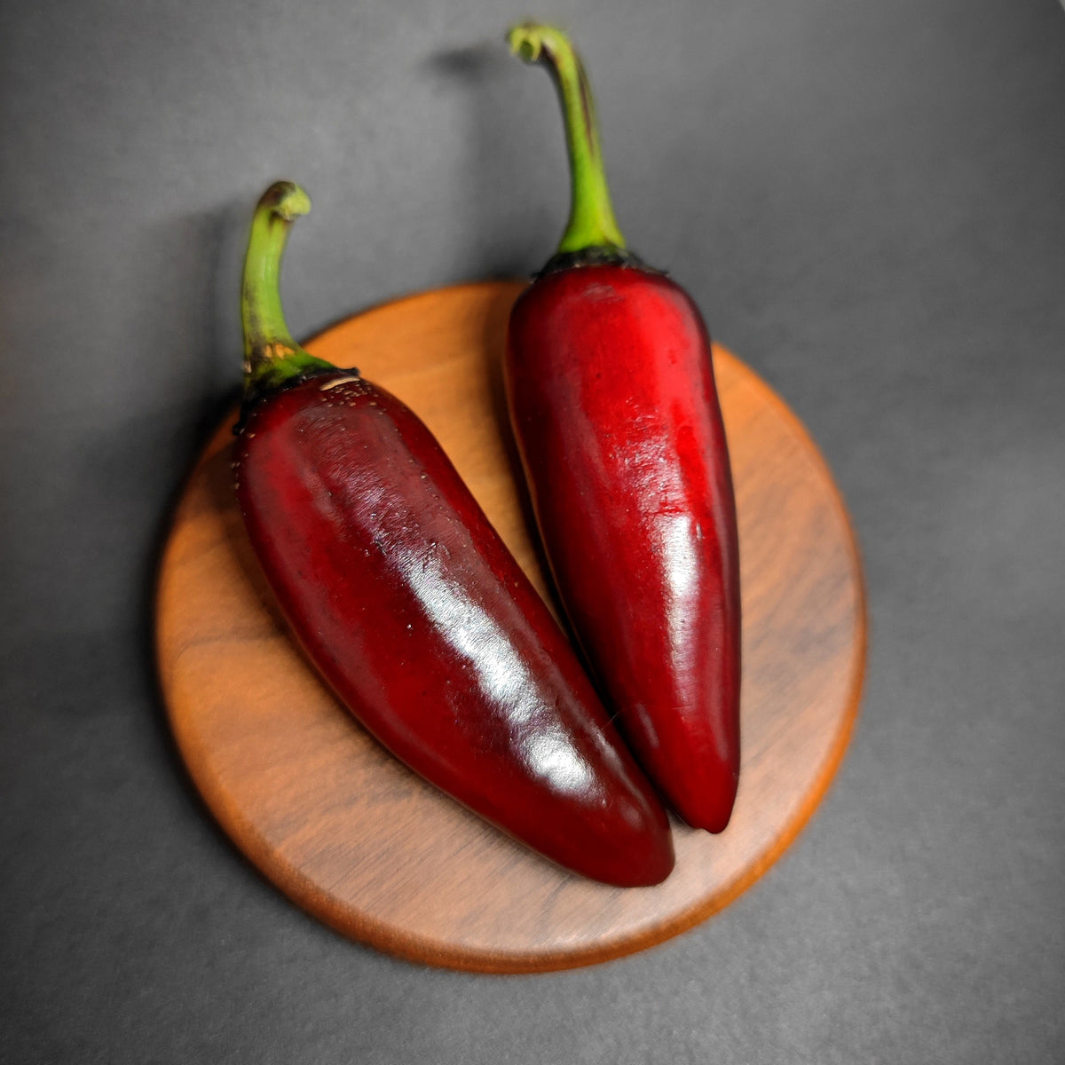 Two Ruby Jalapeno peppers, distinguished by their dark red color and green stems, from PepperMerchant.net rest on a round wooden coaster against a black background.