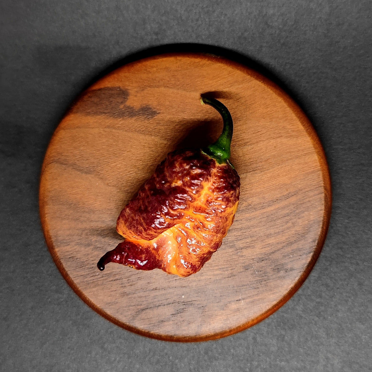 A wrinkled, dark red Carolina Reaper pepper used in a Reaper Sbagliato with a green stem resting on a circular wooden board.