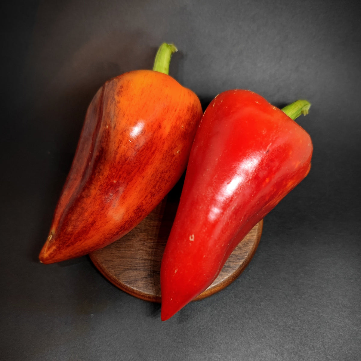 Two Violet Sparkle thick-walled chili peppers, one orange and streaked and one solid red, resting on a small wooden coaster.