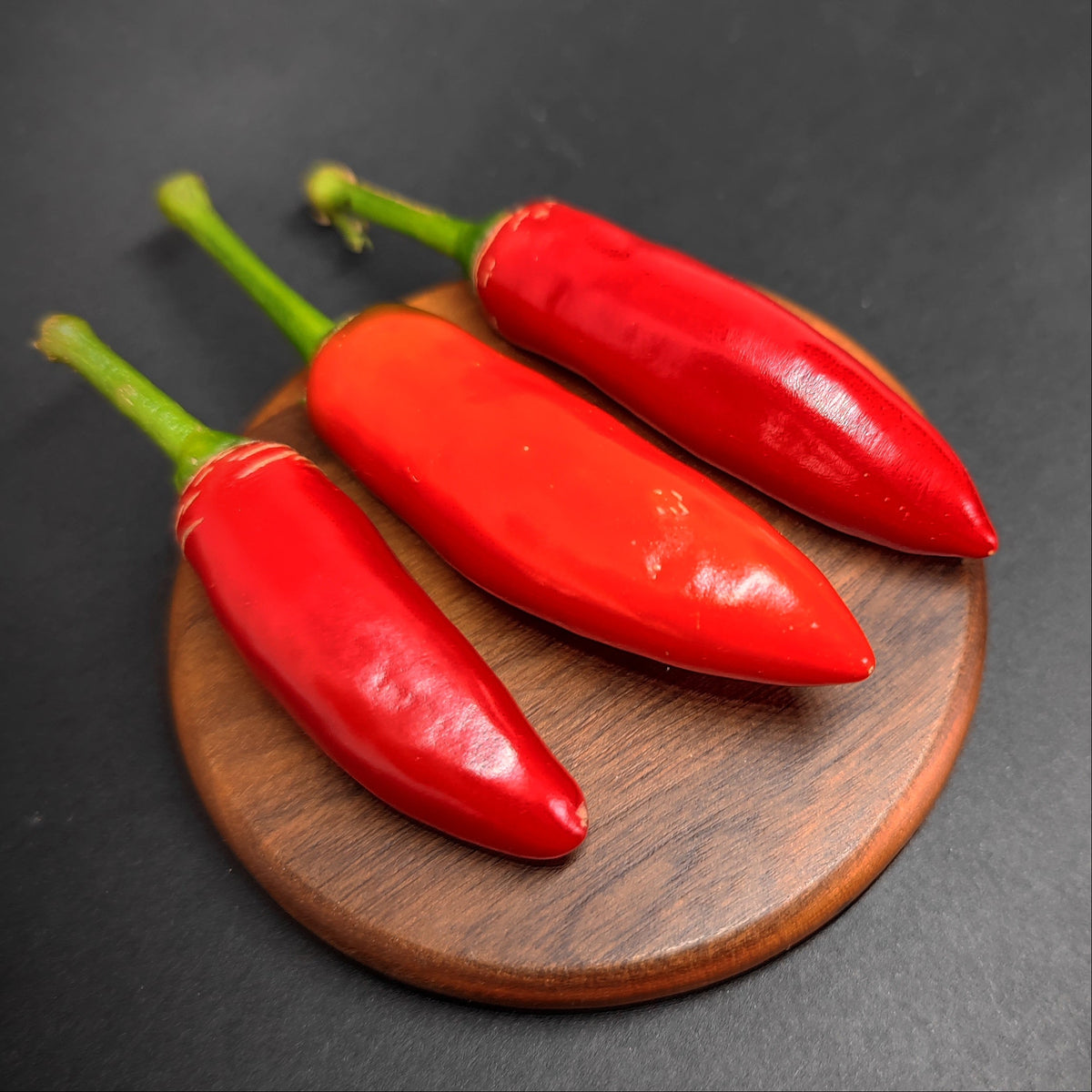 Three red Calabrian peppers from PepperMerchant.net on a small round wooden board.