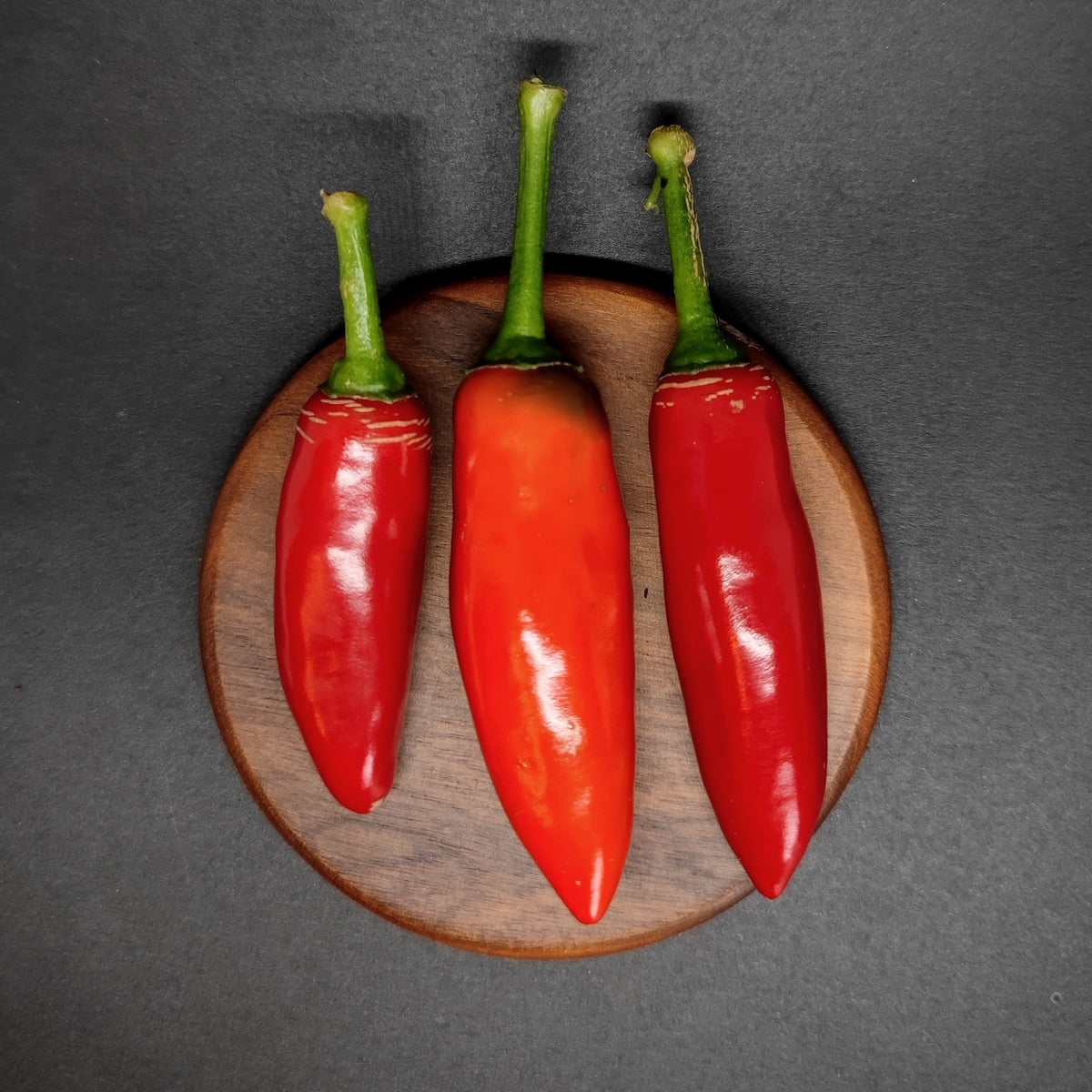 Three red Calabrian peppers with green stems arranged on a circular wooden coaster.