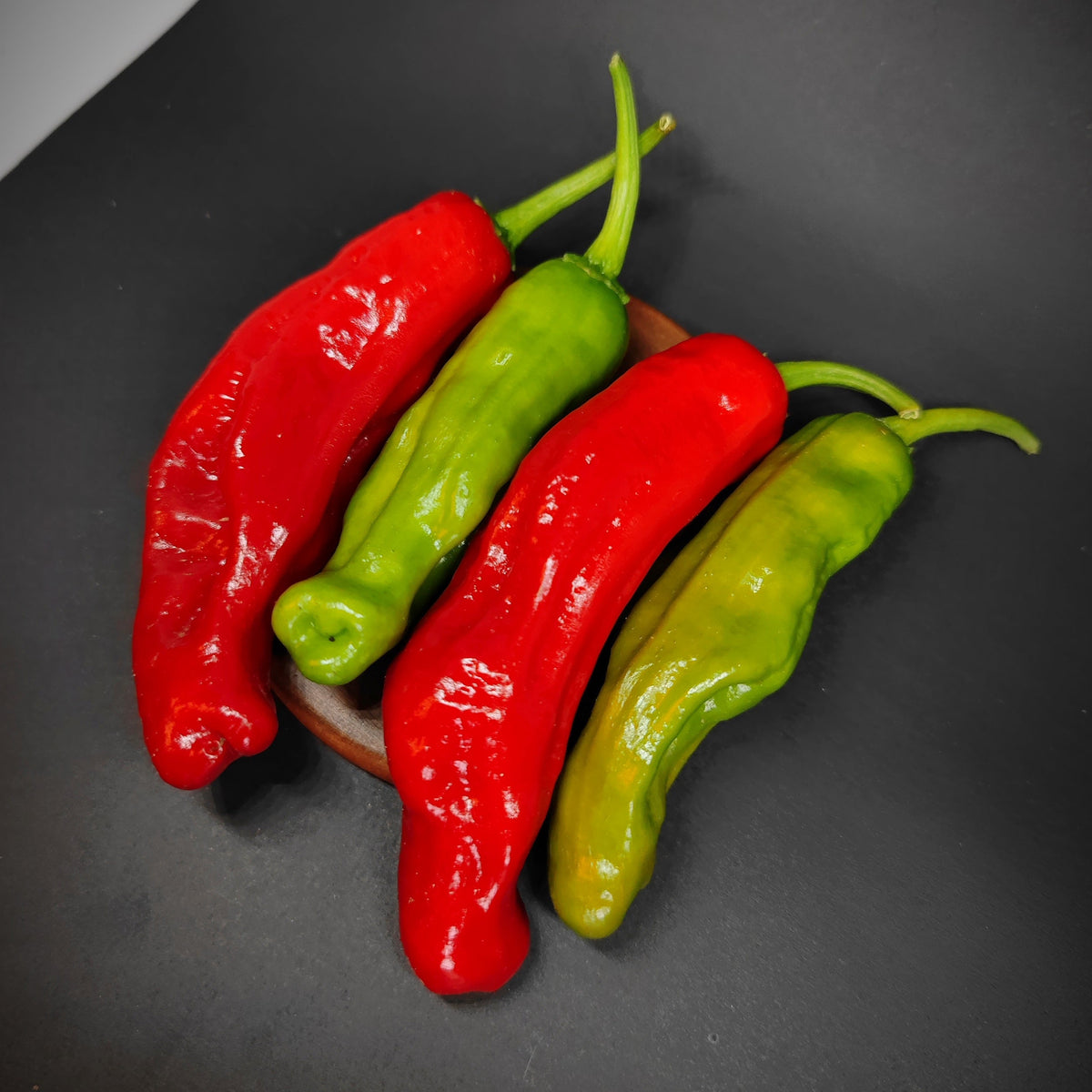 Four long Shishito peppers from PepperMerchant.net, two red and two green, are arranged side by side on a small wooden tray against a dark background, evoking the vibrant appearance of this beloved Japanese pepper variety known for its mild heat.
