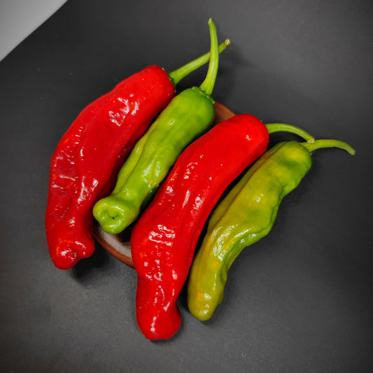 Four long Shishito peppers from PepperMerchant.net, two red and two green, are arranged side by side on a small wooden tray against a dark background, evoking the vibrant appearance of this beloved Japanese pepper variety known for its mild heat.
