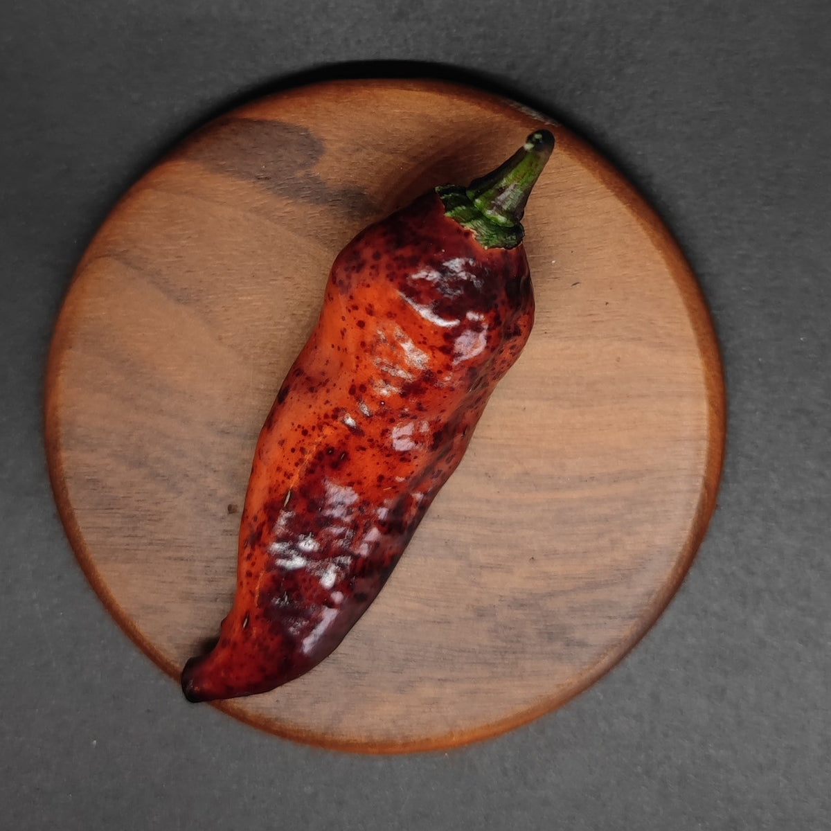 A dark ruby Dark Ruby pepper with speckled skin resting on a small wooden circular board.